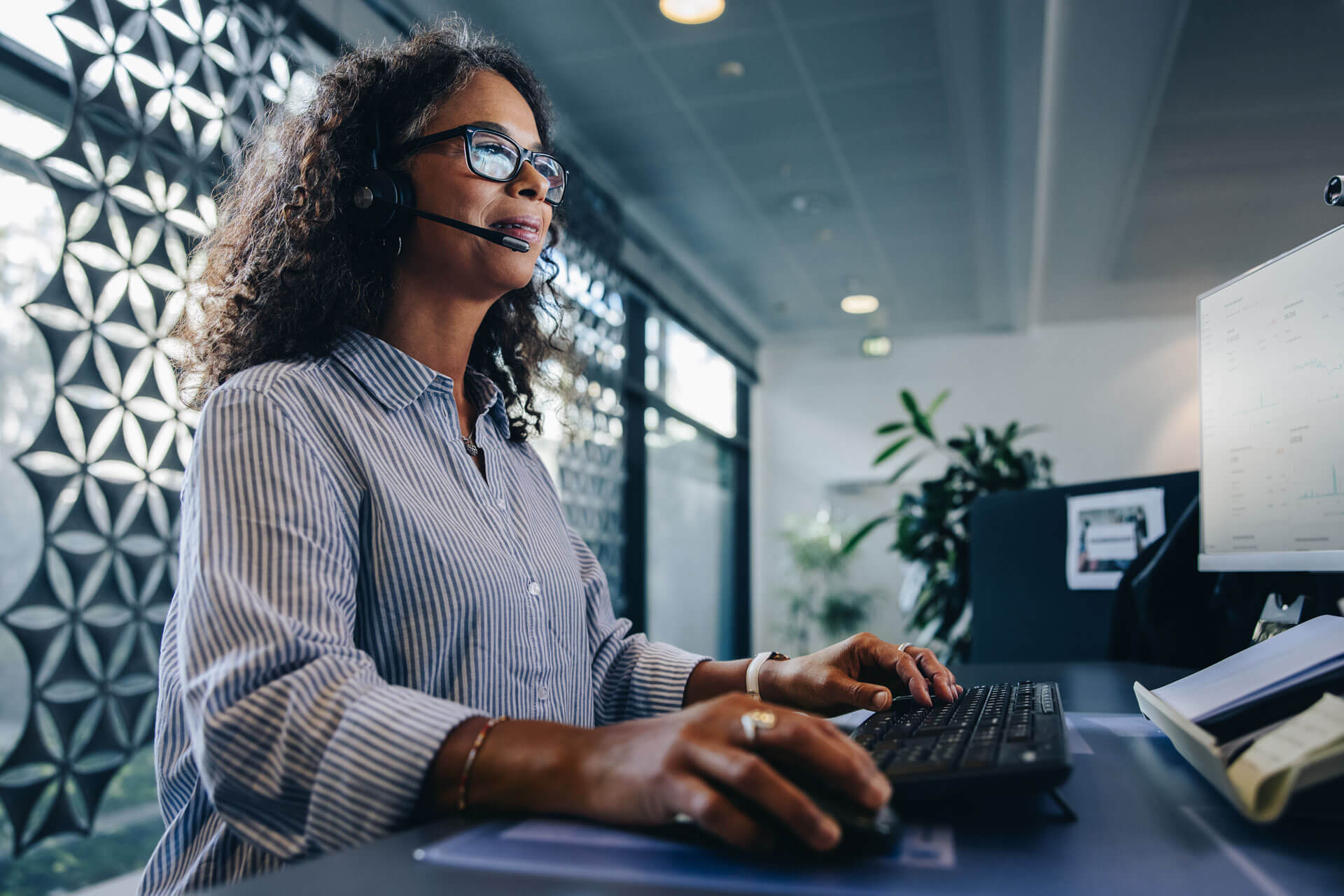 A woman with curly hair and glasses, wearing a striped shirt and headset, types on a keyboard in a modern office to represent Cogency Global’s national registered agent and registered office services.