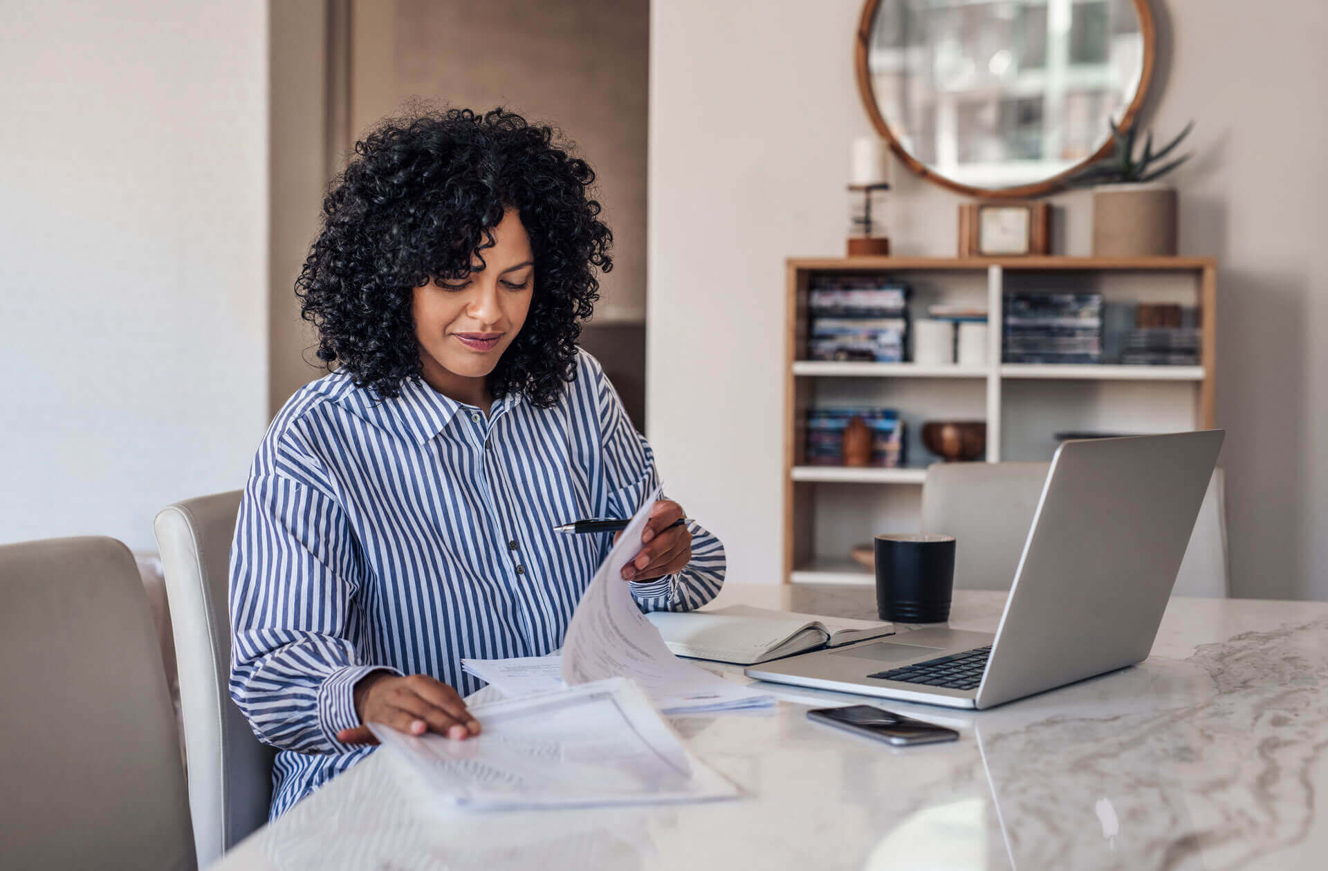 A woman with curly hair wearing a striped shirt is sitting at a table with a laptop, a cup, and some documents. She is holding a pen and looking at the papers in front of her. A bookshelf and a round mirror are in the background.