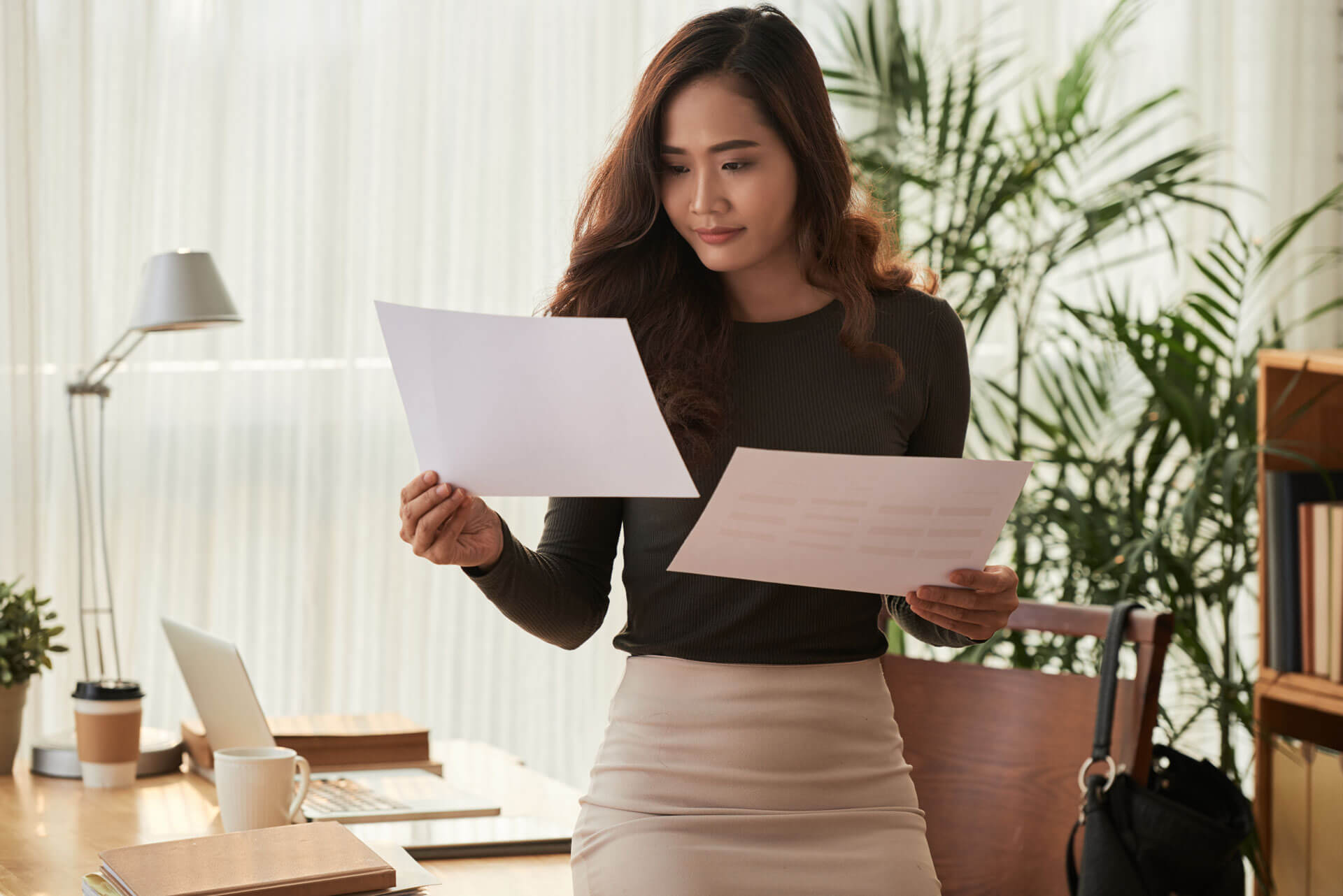 A woman in a business setting is standing and reviewing two documents. She wears a dark green top and a beige skirt. In the background, there is a desk with a laptop, a cup, books, and various office plants.