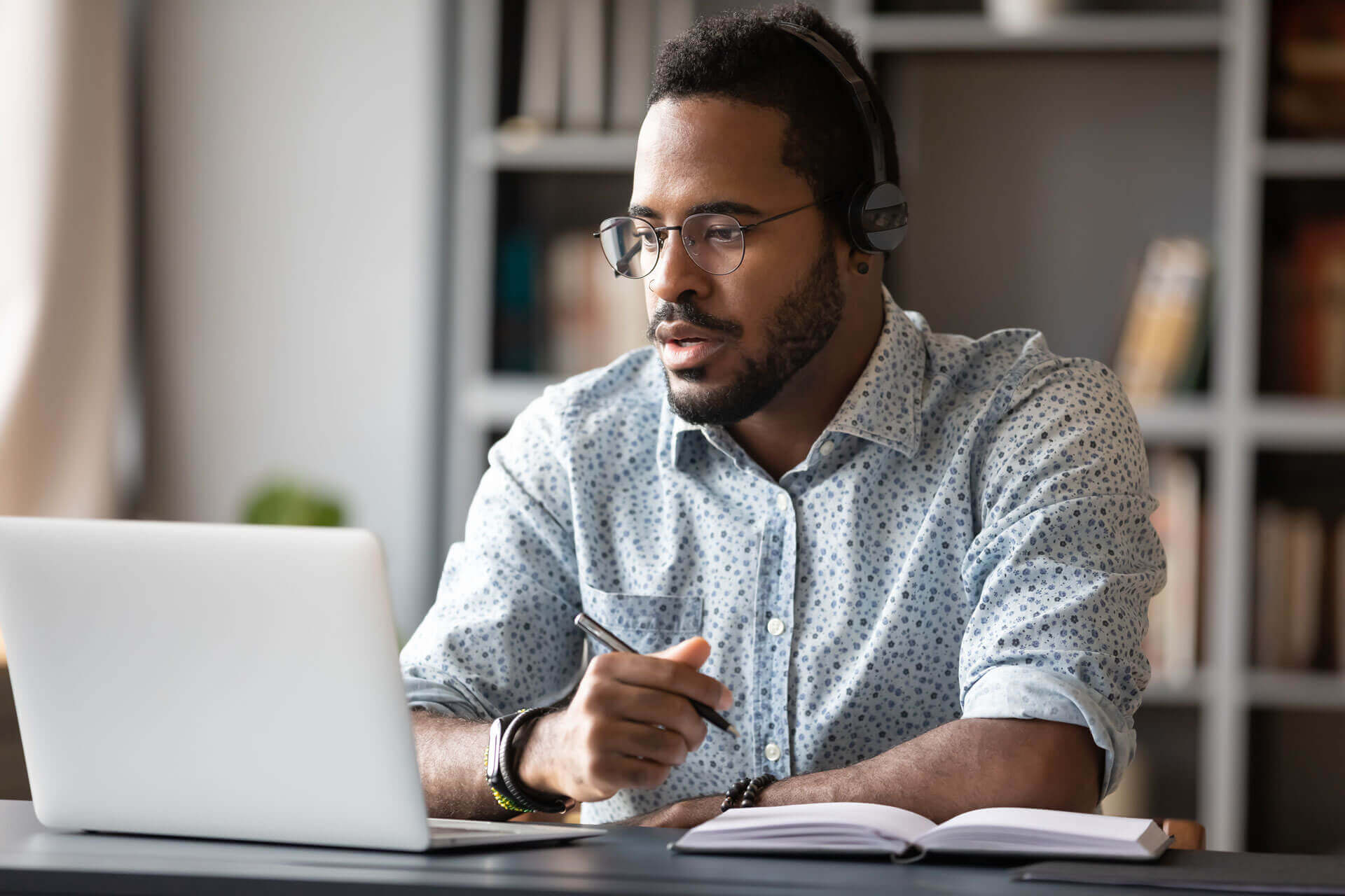 A man with glasses and headphones, wearing a patterned button-up shirt, is sitting at a desk working on a laptop. He is holding a pen and a notebook is open in front of him. Shelves with books and decor are visible in the background.