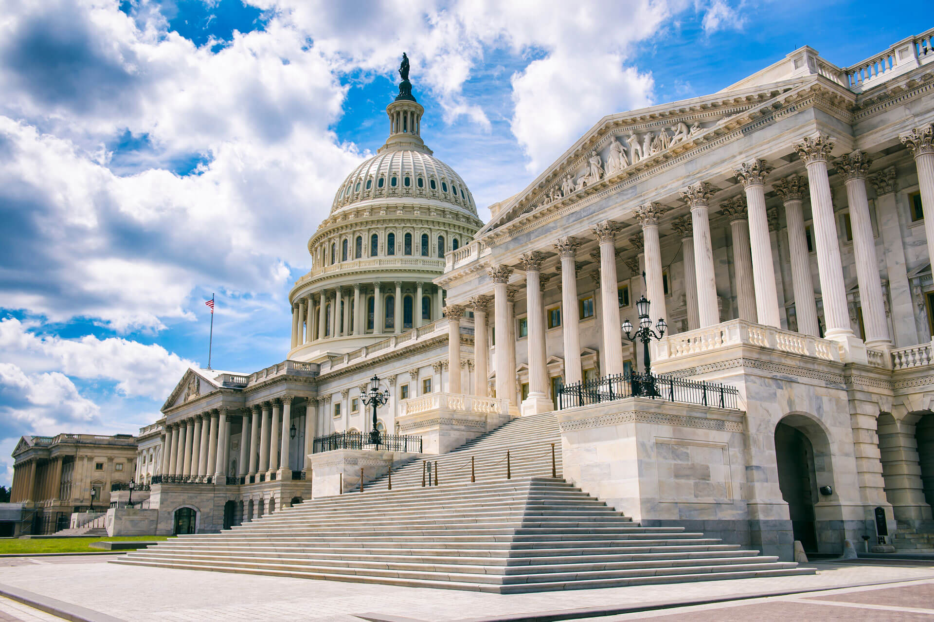 The United States Capitol building in Washington, D.C., is shown with its large dome, classical columns, and steps leading up to the entrance. The sky is bright with white clouds scattered against a blue backdrop.