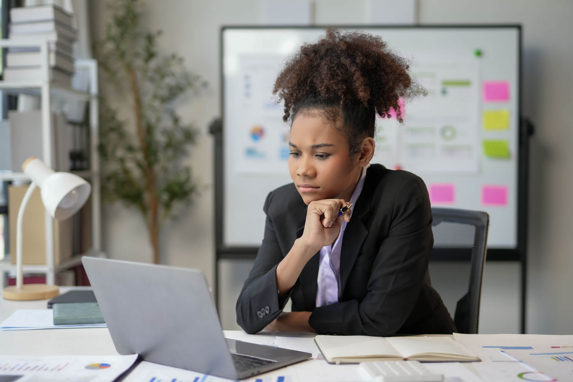 A person in a business suit is sitting at a desk, focused on a laptop screen. There are documents, a notebook, and office supplies on the desk. A whiteboard with charts and graphs is in the background, along with a potted plant and shelves with books and files.