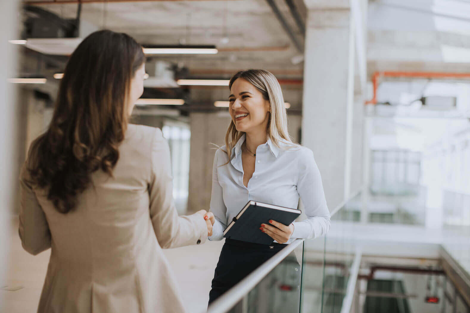 Two women in business attire shake hands and smile in a modern office near a glass railing, engaged in a positive conversation, representing the emphasis Cogency Global's places on creating the best experience for UCC search and lien search clients.