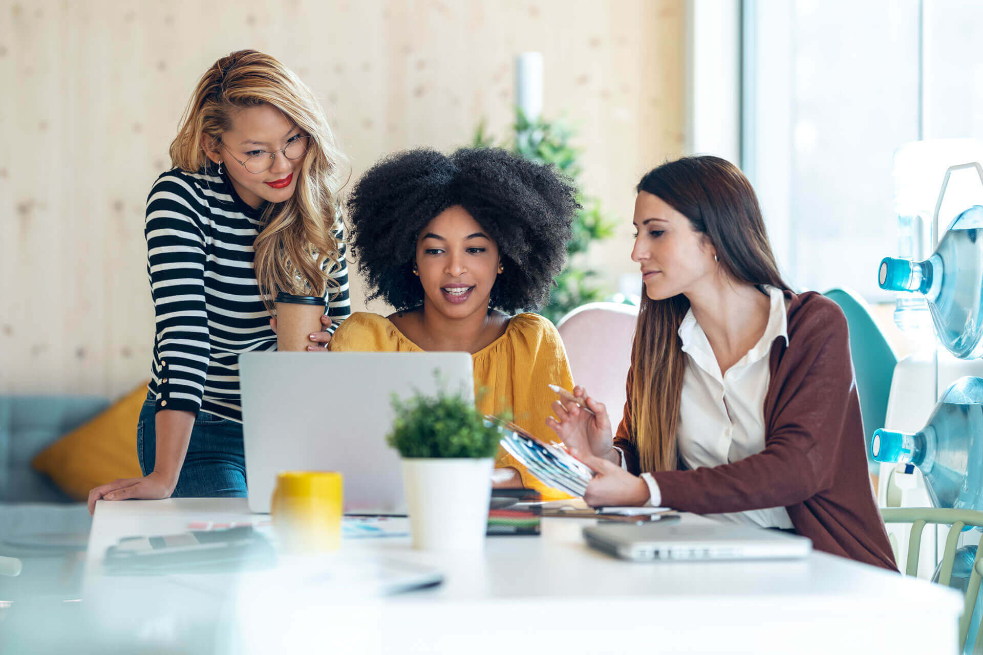 Three women collaborate at a desk in a modern office, focused on a laptop, likely working on UCC filing with Cogency Global's industry-leading software UCC ProFile.