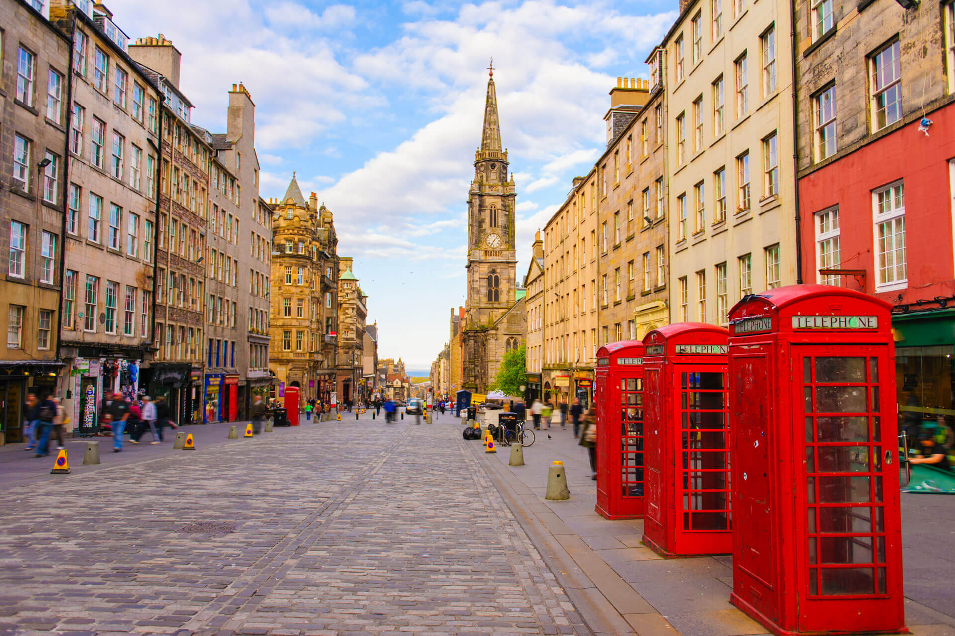 A cobblestone street in a historic city, lined with old buildings. Red telephone boxes are on the right. A tall, ornate spire is in the distance under a blue sky with clouds. People walk along the sides of the street.