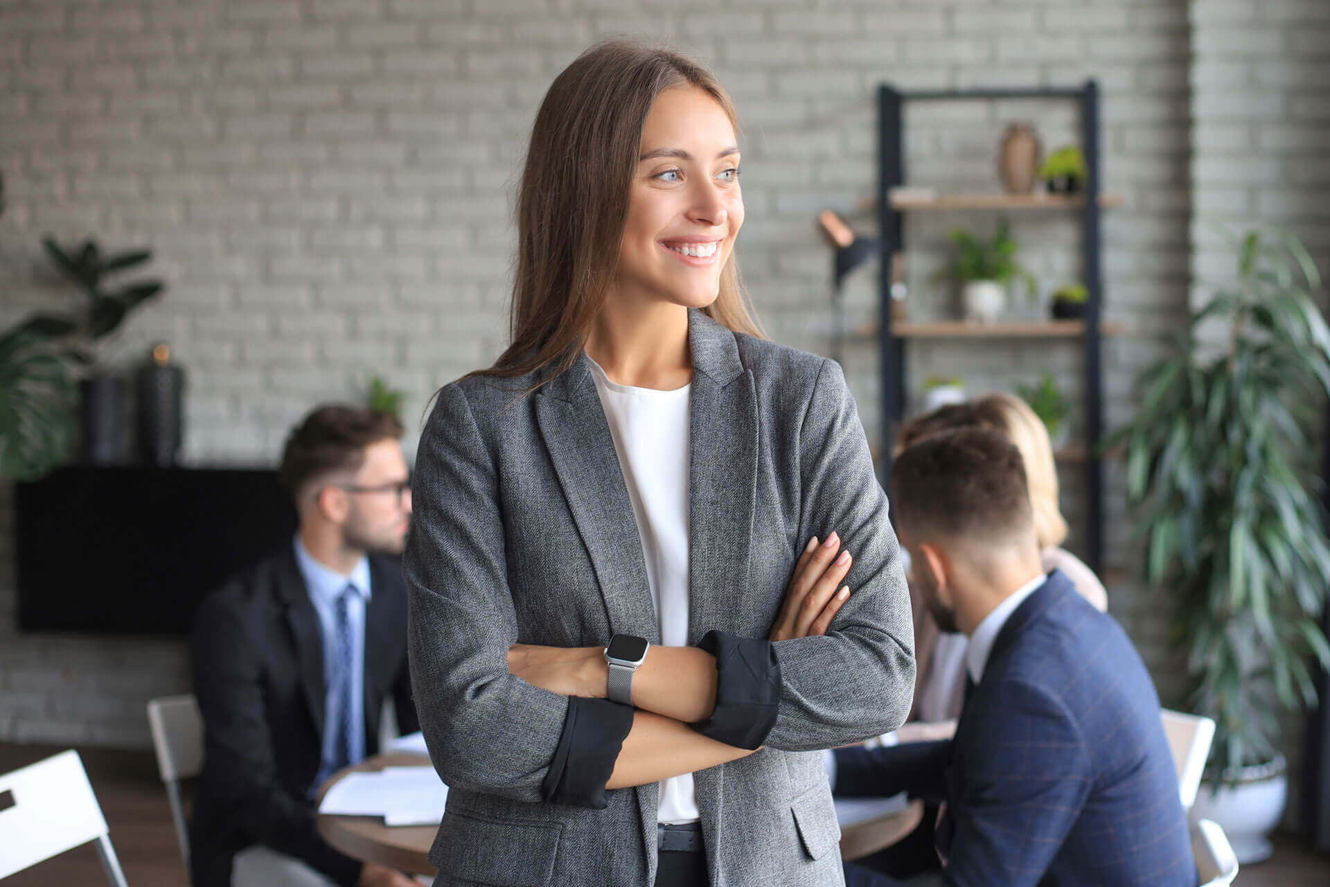 A smiling woman in a grey blazer stands with her arms crossed in an office setting. In the background, three colleagues are engaged in a discussion around a table. The room features a brick wall, various plants, and shelves with decorative items.
