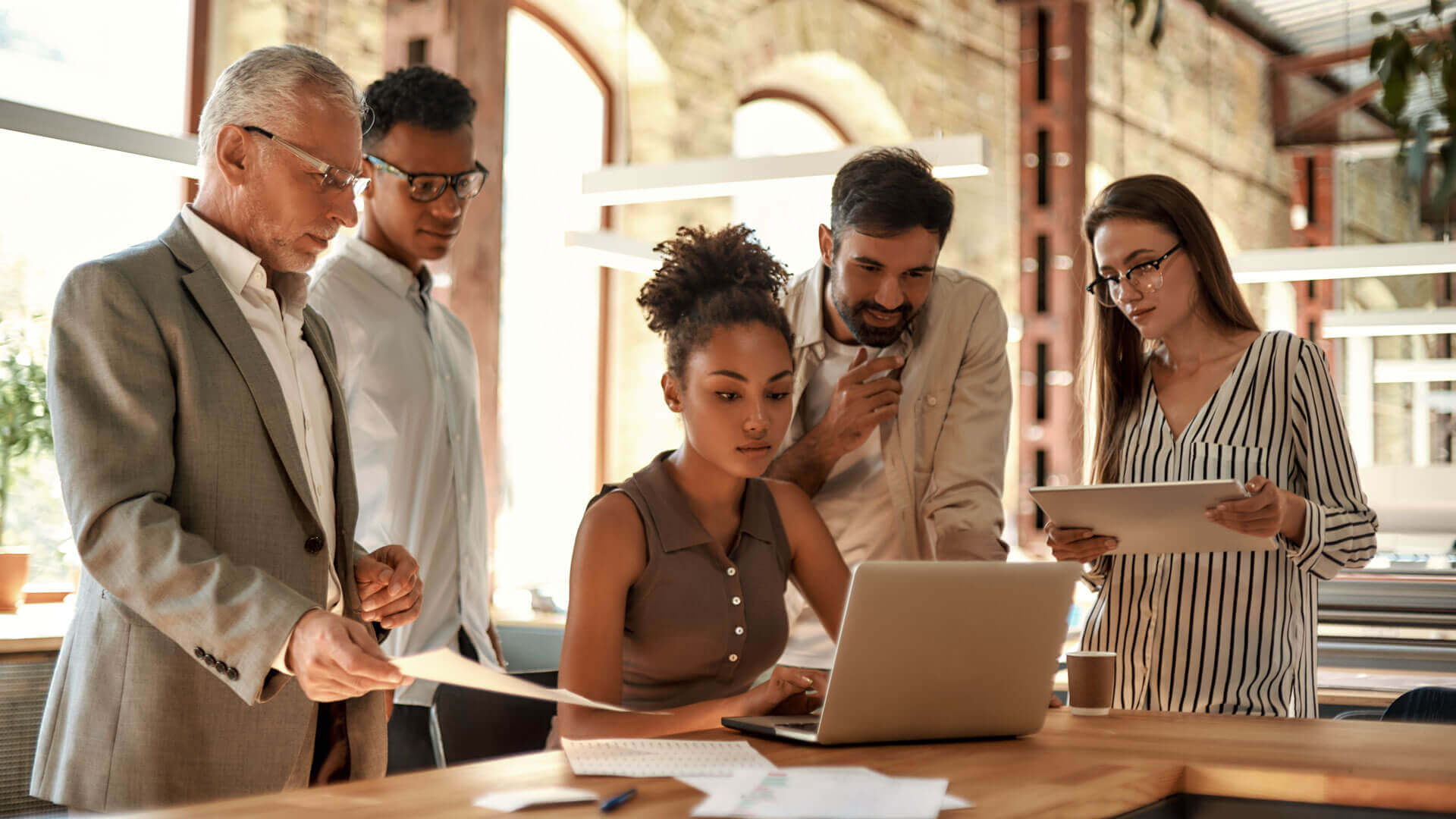 A diverse group of five coworkers engaged in a discussion in a modern office. A woman using a laptop sits at a desk in the center, while two men and two women stand around her holding papers and tablets, collaborating attentively.