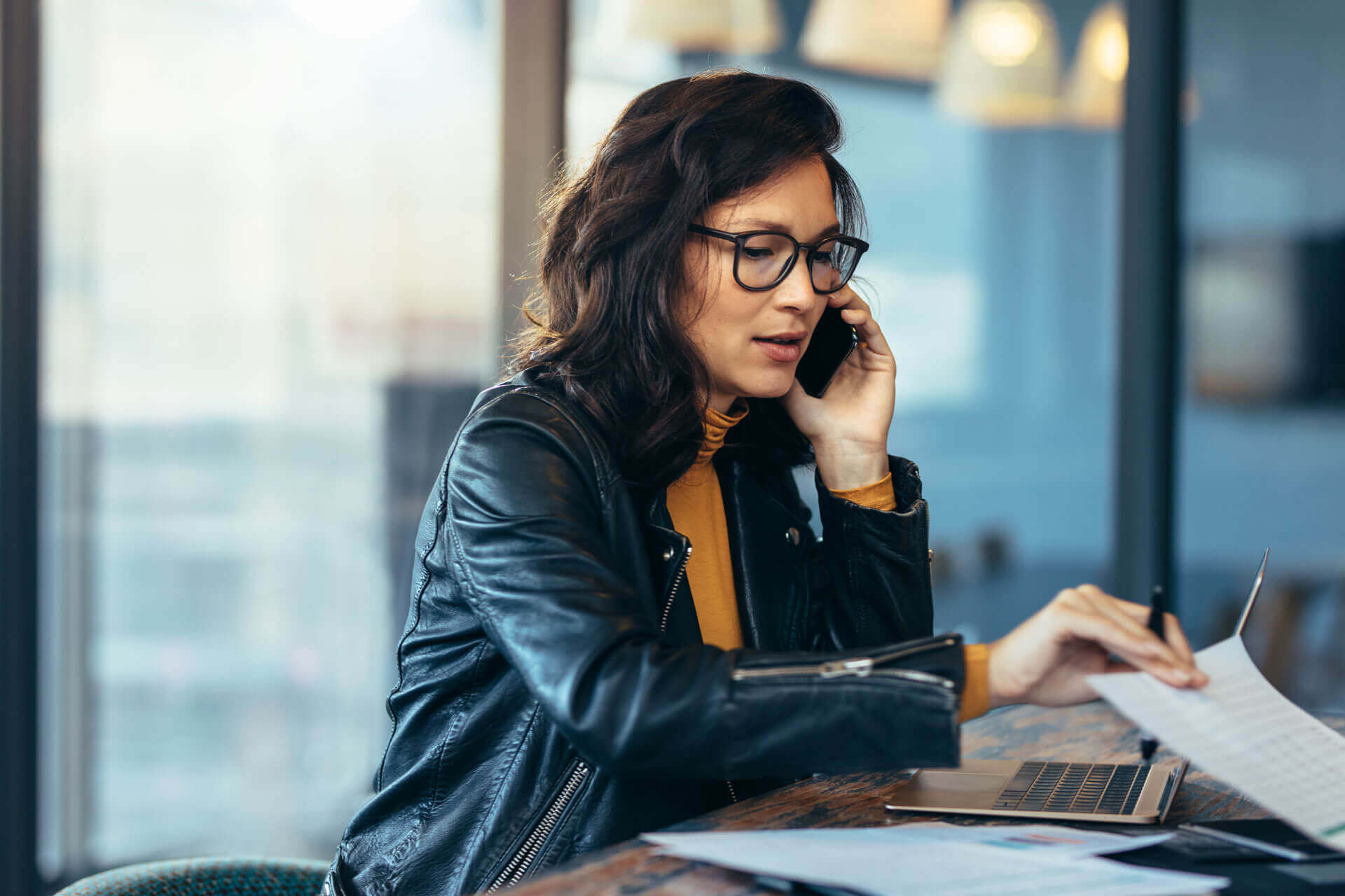 A woman with glasses and a black leather jacket is talking on the phone while looking at documents. She is seated at a desk with a laptop and papers in front of her in a modern, well-lit workspace.