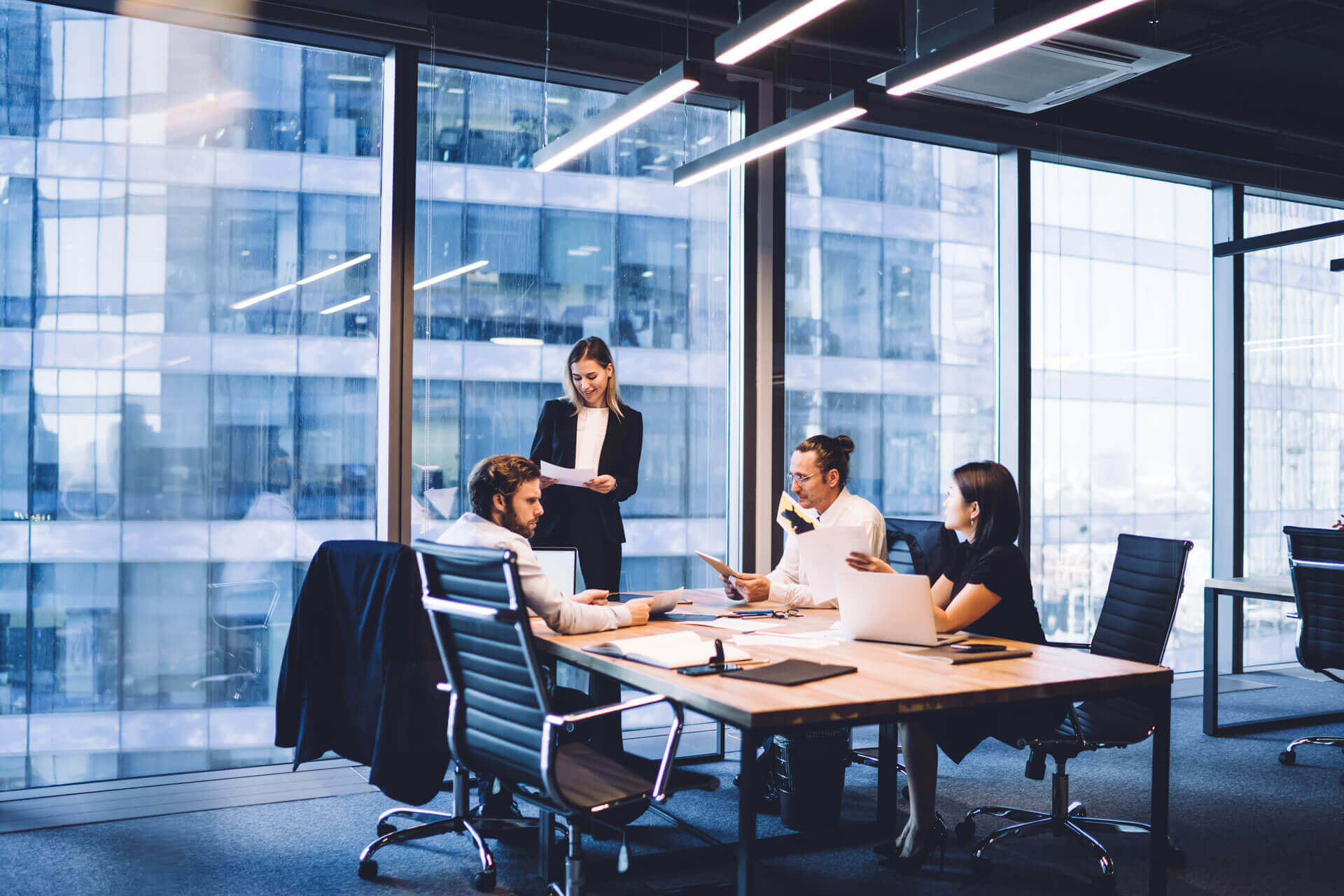 Four people are in a modern office with large windows, seated around a conference table. Three are sitting, engaged in discussion and reviewing documents, while one stands, presenting information. Outside, a tall building can be seen.