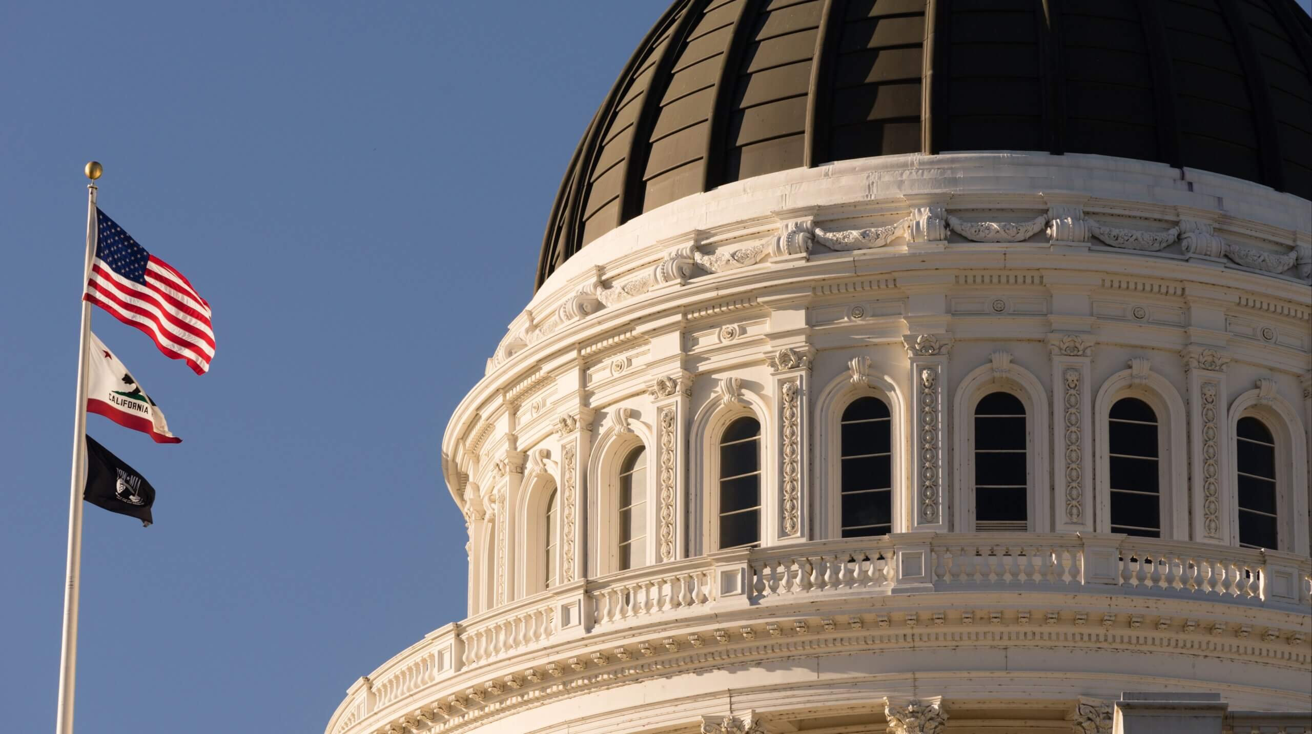 Close-up view of a white domed building with arched windows, likely a capitol building. An American flag, a state flag, and a third flag are flying on a pole against a clear blue sky.