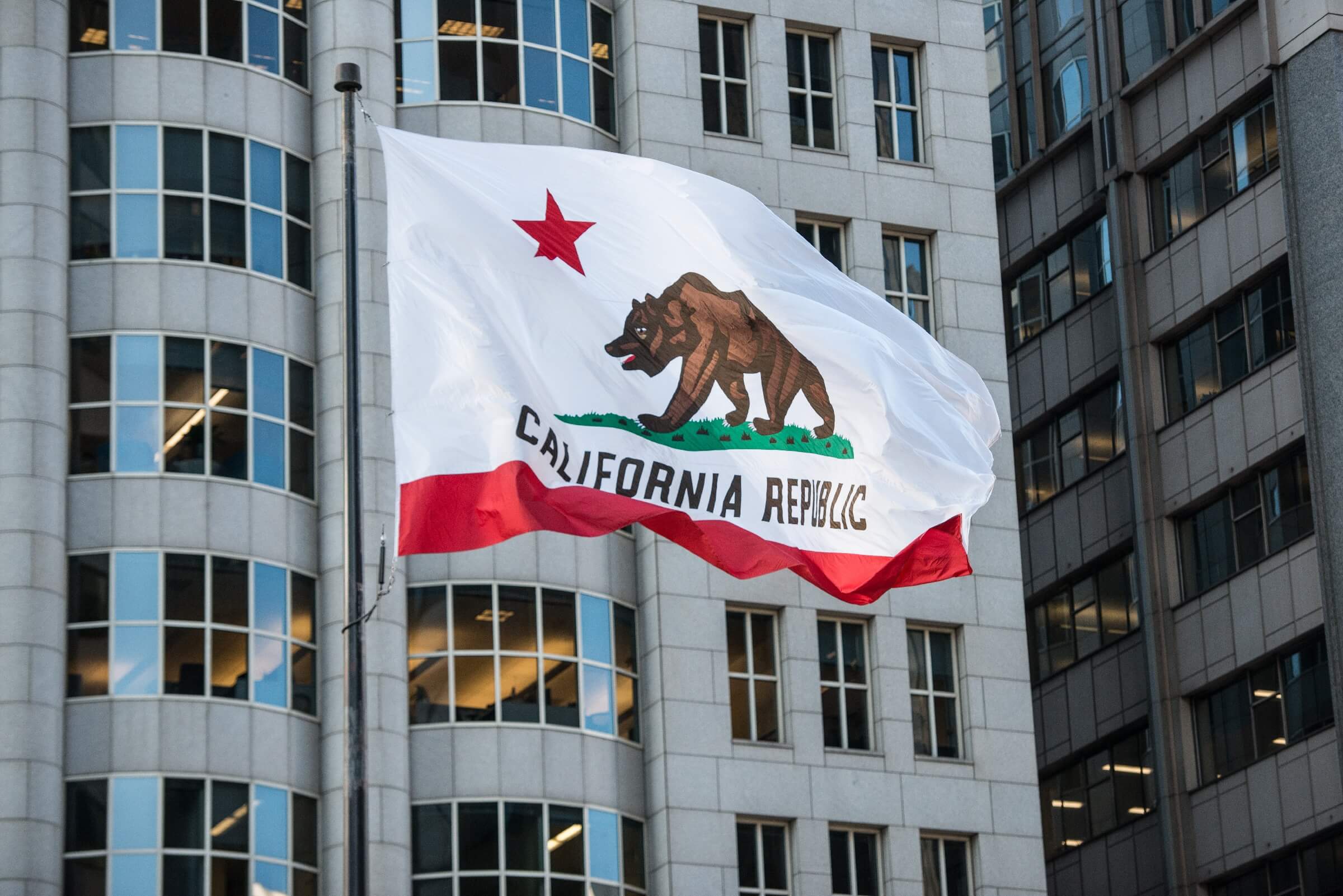 The California state flag with a bear and star waves in front of a modern building with large windows.