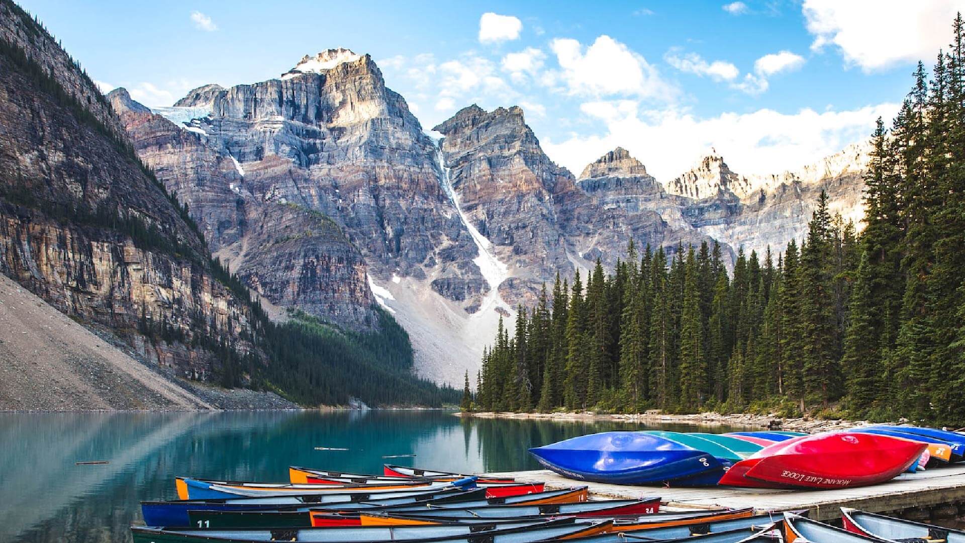 A scenic view of colorful canoes lined up on the shore of a calm mountain lake. In the background, tall pine trees and rugged, snow-capped mountains under a partly cloudy sky. The water reflects the mountains and trees.