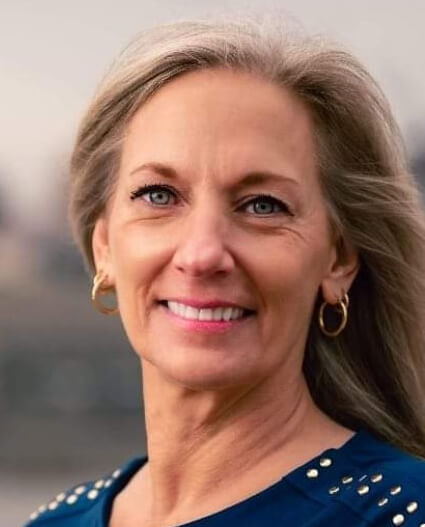 A woman with long gray hair smiles while standing outdoors. She is wearing a blue top with decorative elements on the shoulders. A blurred cityscape is visible in the background.