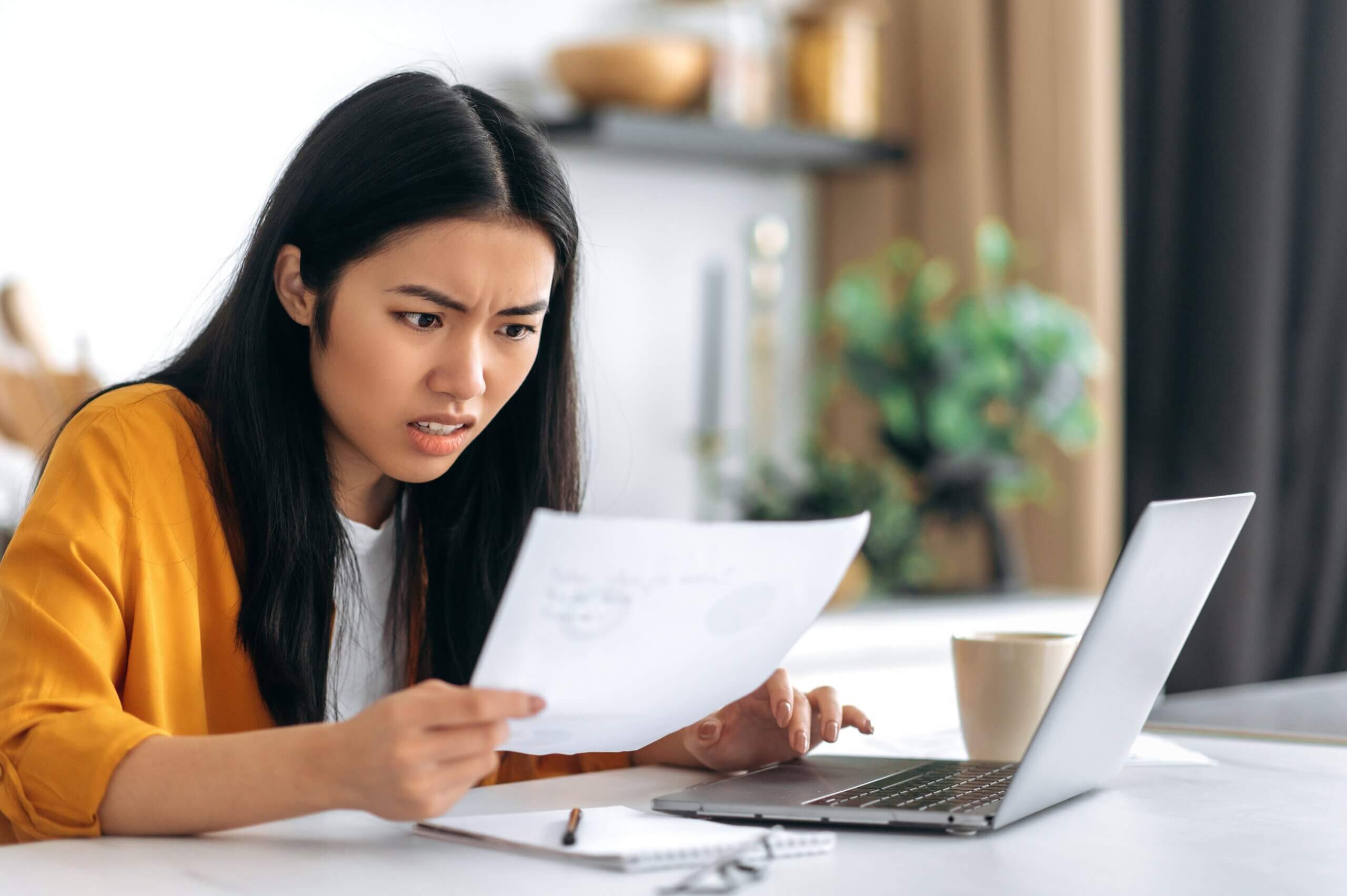 A woman in a yellow cardigan appears confused or concerned while looking at a document in her hand. She sits at a table with a laptop, notepad, and a coffee cup. The background shows shelves with decor.
