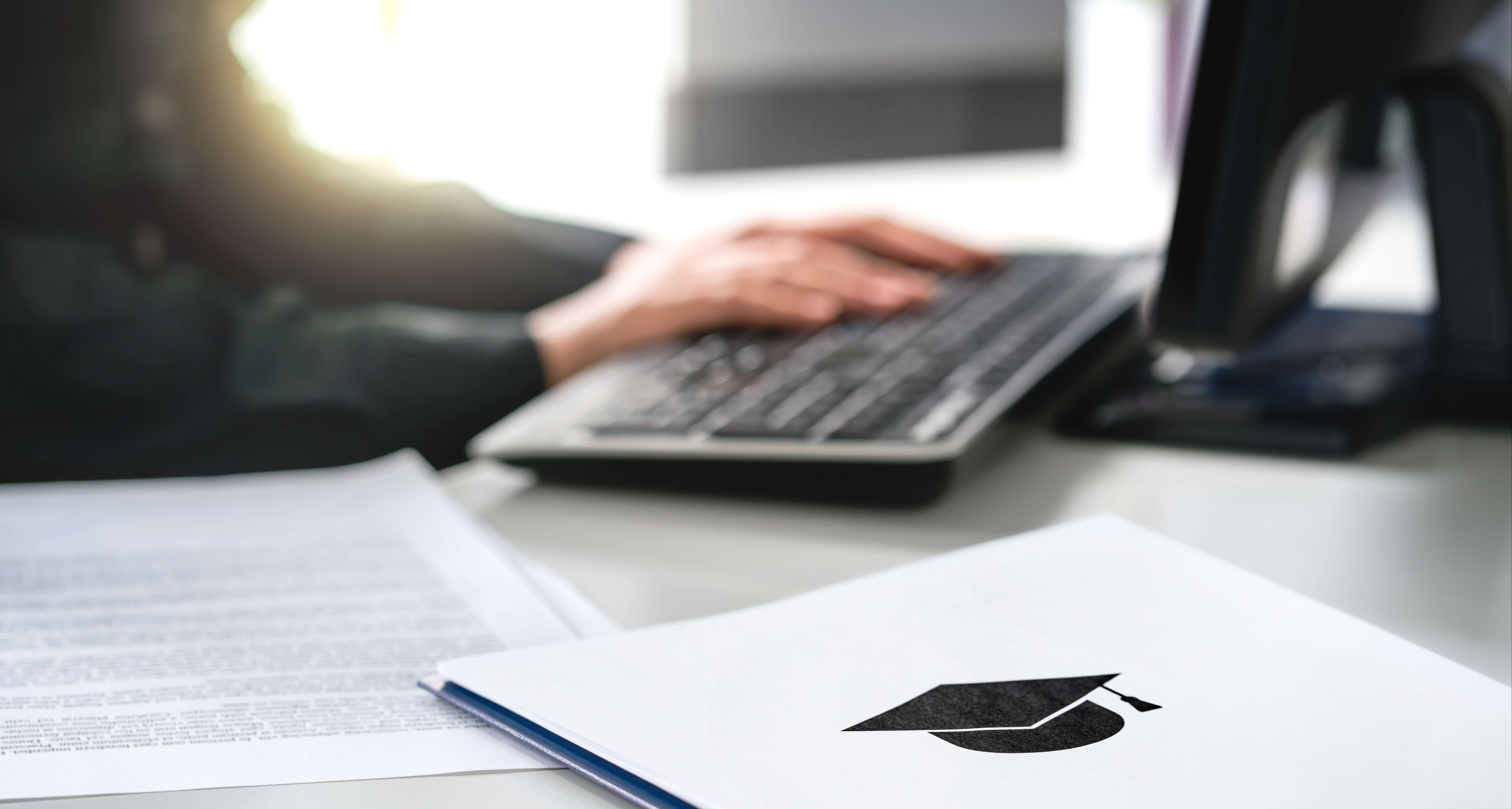 Person typing on a keyboard with a graduation cap symbol on a document in the foreground. Sunlight is streaming in, illuminating the workspace.