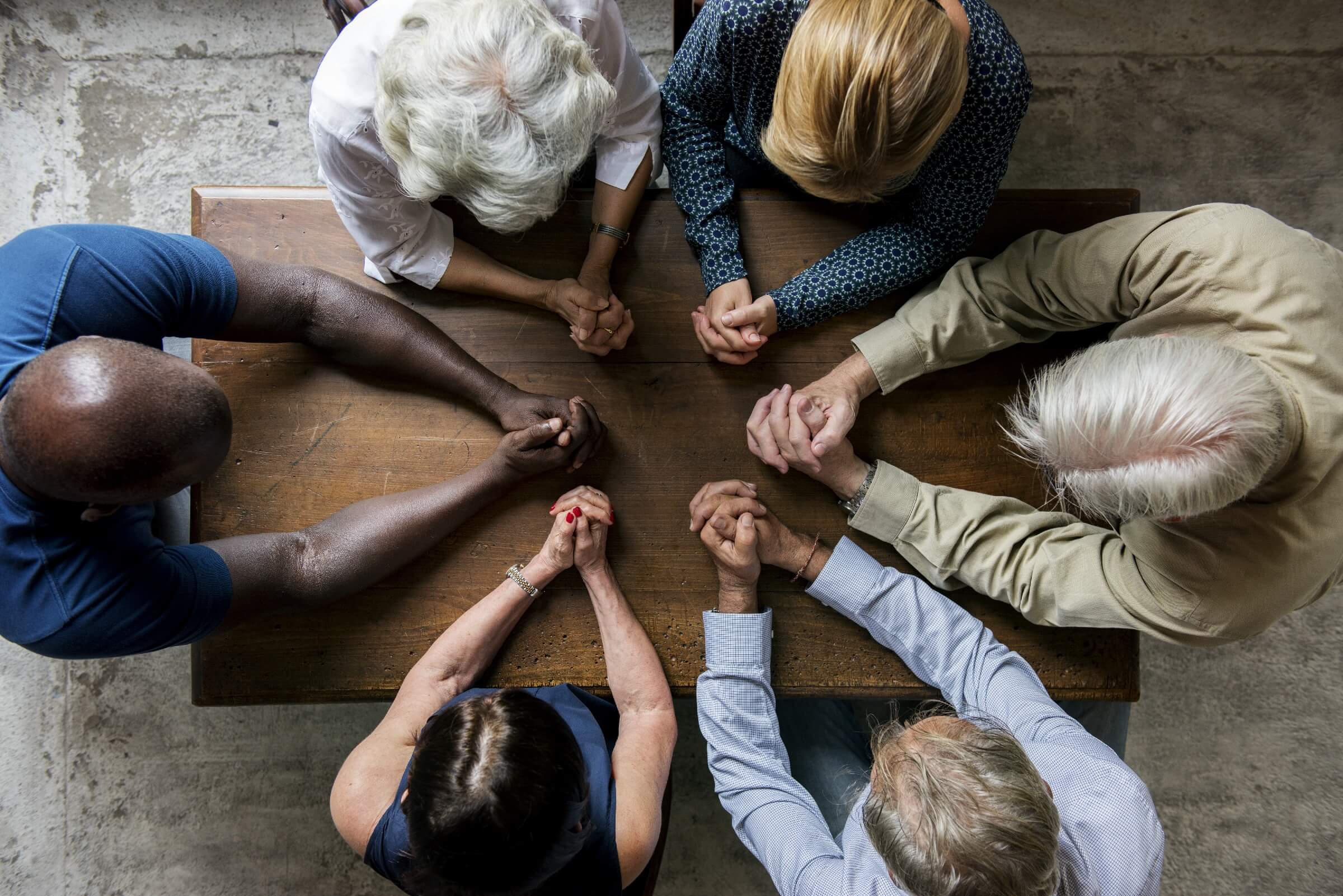 A diverse group of six people sitting around a wooden table, viewed from above. They are holding hands, suggesting unity or prayer. The individuals vary in age and ethnicity, with their focus on the center of the table.