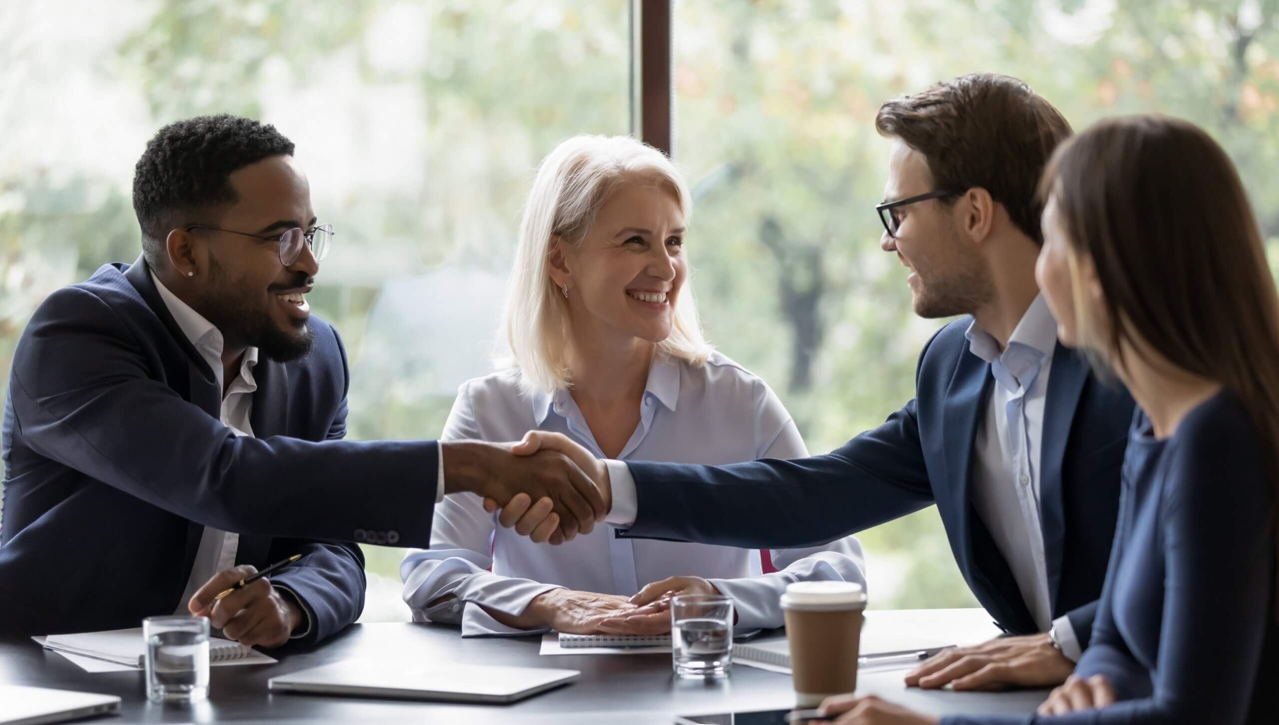 Four people in business attire sit at a table, smiling. Two are shaking hands across the table, with laptops and a coffee cup in front. A large window with greenery outside is in the background.