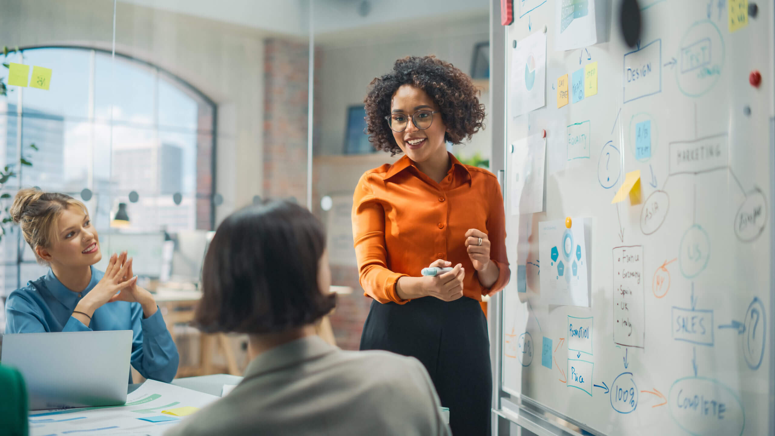 A woman in an orange blouse presents ideas on a whiteboard to colleagues in a modern office. Two women sit listening; one claps. The whiteboard is filled with colorful notes and charts. The room is bright with a large window in the background.