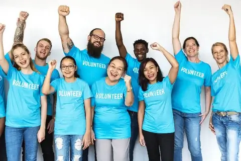 A diverse group of people wearing blue "VOLUNTEER" t-shirts stand together cheerfully with raised fists, smiling and celebrating. They are in front of a plain white background.