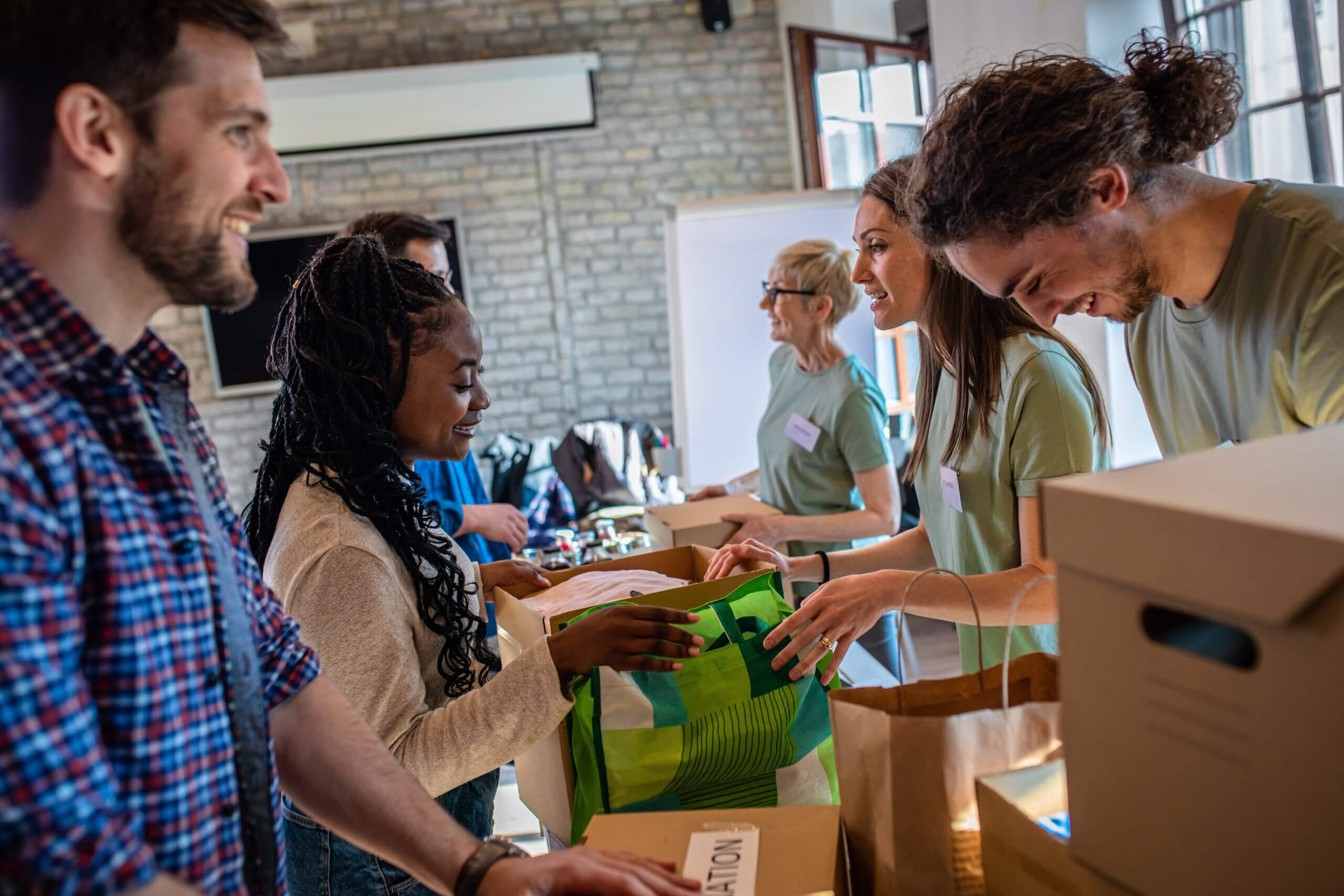 A diverse group of people are joyfully packing boxes and bags with supplies, likely volunteering at a community center. They are indoors, surrounded by cardboard boxes, and everyone seems engaged and happy.