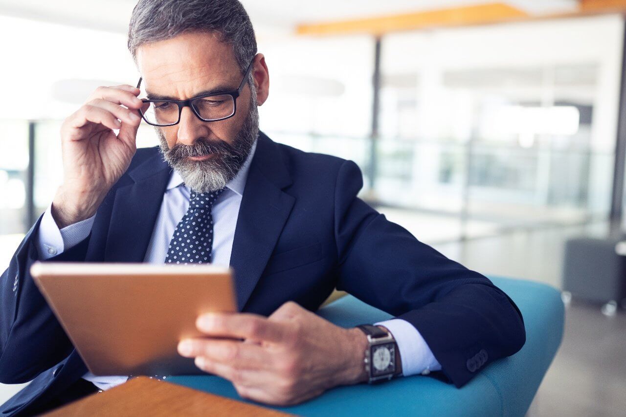 A bearded man in a suit and glasses is sitting on a blue chair, holding an electronic tablet with one hand and adjusting his glasses with the other. He is focused on the tablet screen in a modern office setting.