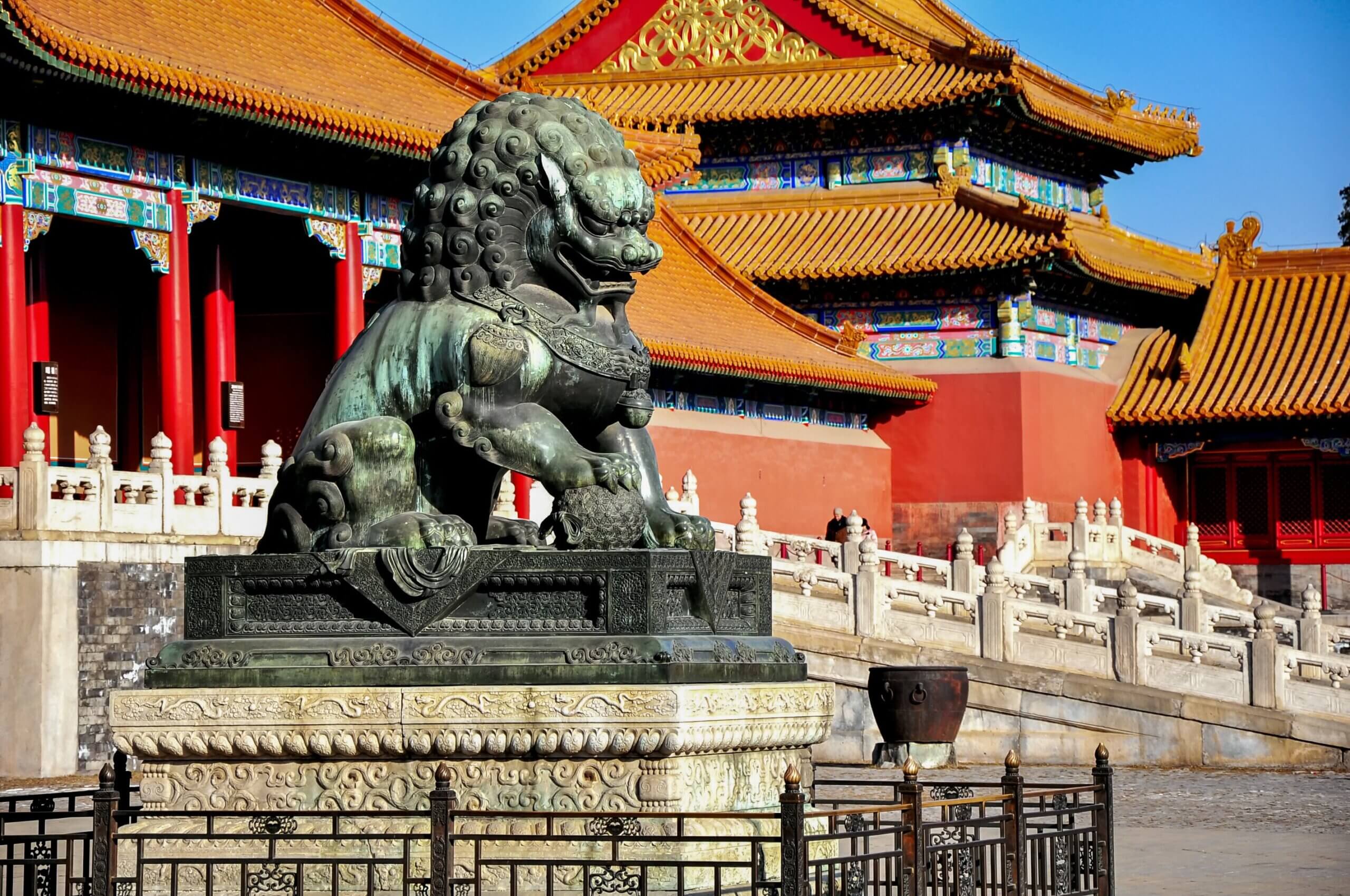 A bronze lion statue stands before an ornate building in the Forbidden City, Beijing. The building features red walls, gold-tiled roofs, and intricate detailing. Stone steps and ornate railings are visible under a clear blue sky.