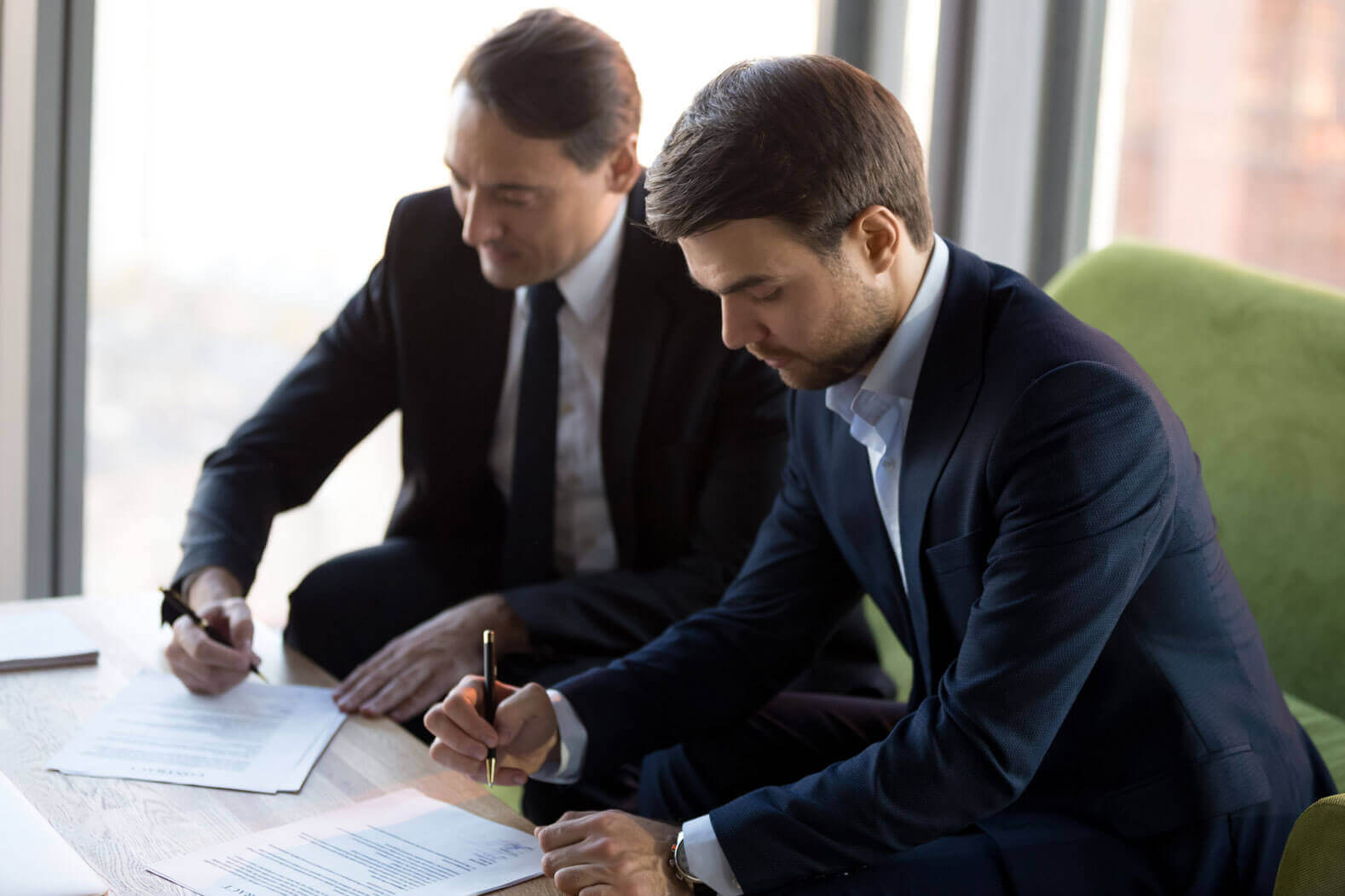 Two professional men in business attire are seated at a table, concentrating as they sign documents. The background features large windows with daylight streaming in, and a green chair is visible beside them. Both are focused on their task, using pens to sign papers.