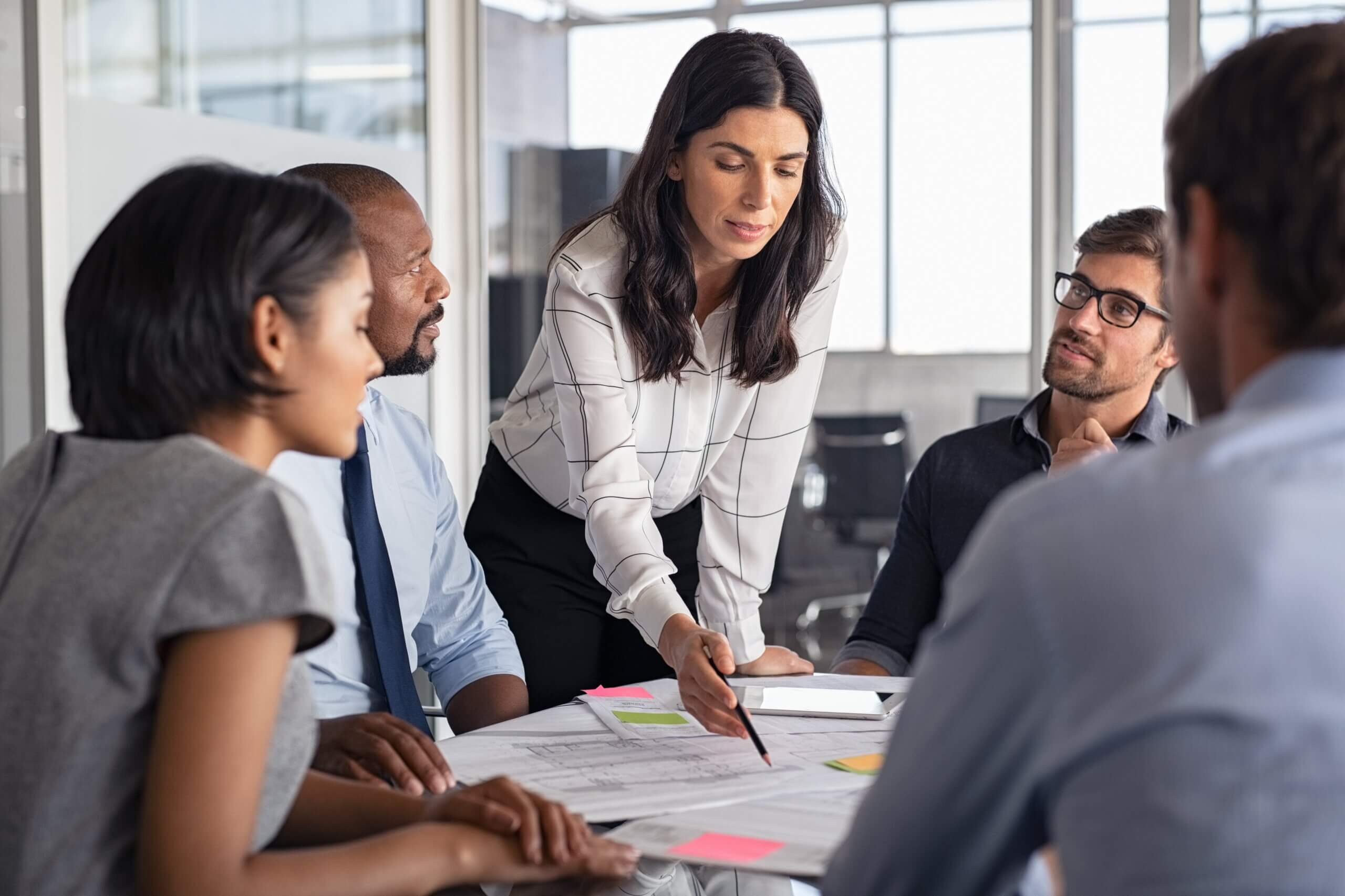 A diverse group of five people in a modern office setting are engaged in a meeting. One woman stands, pointing at documents on the table, while the others listen attentively. The table has papers and sticky notes on it.