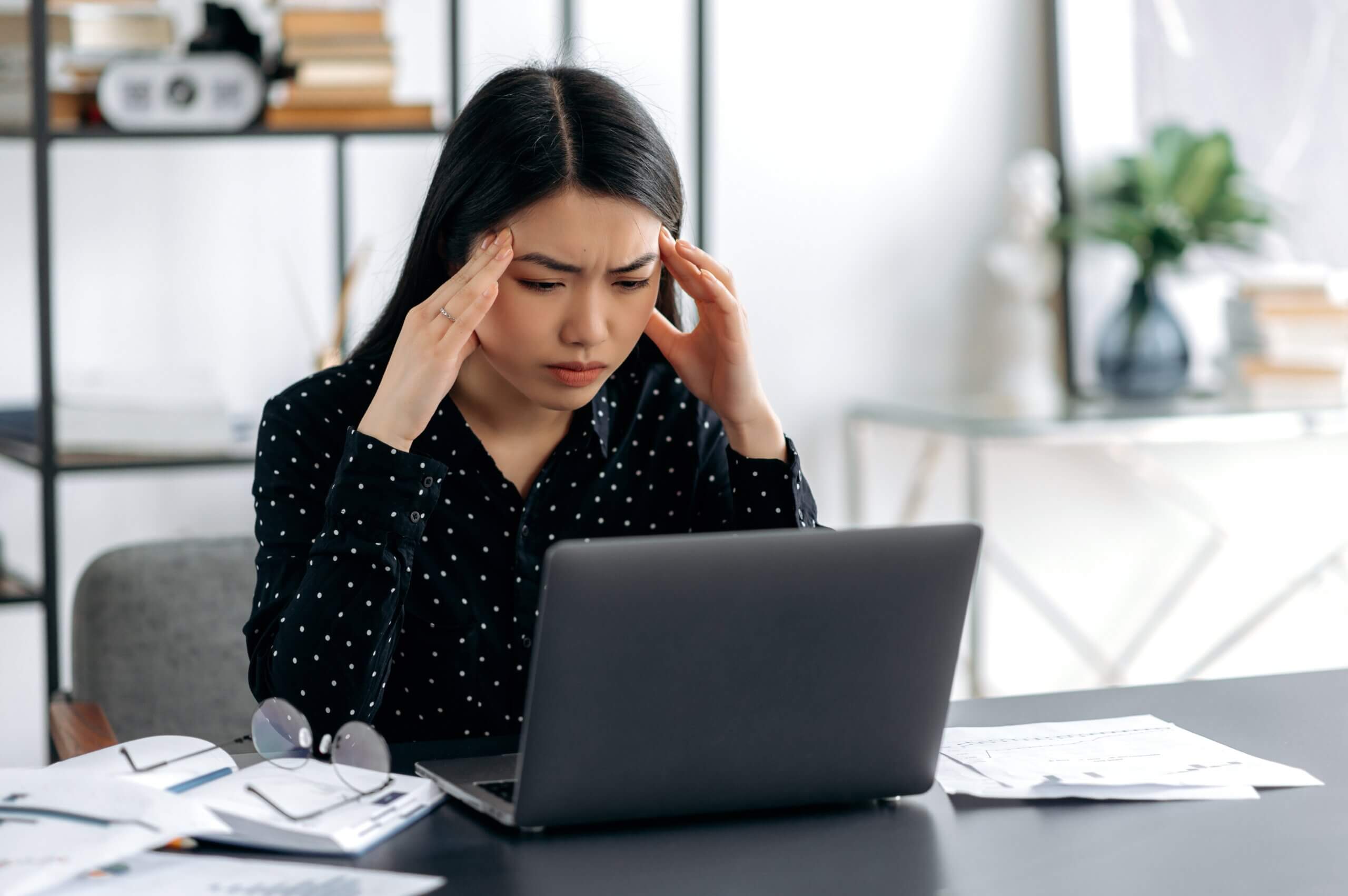 A woman with long dark hair sits at a desk, looking stressed. She holds her temples with her fingers while staring at a laptop. Papers, a pen, and glasses are scattered on the desk. Bookshelves and a plant are in the background.