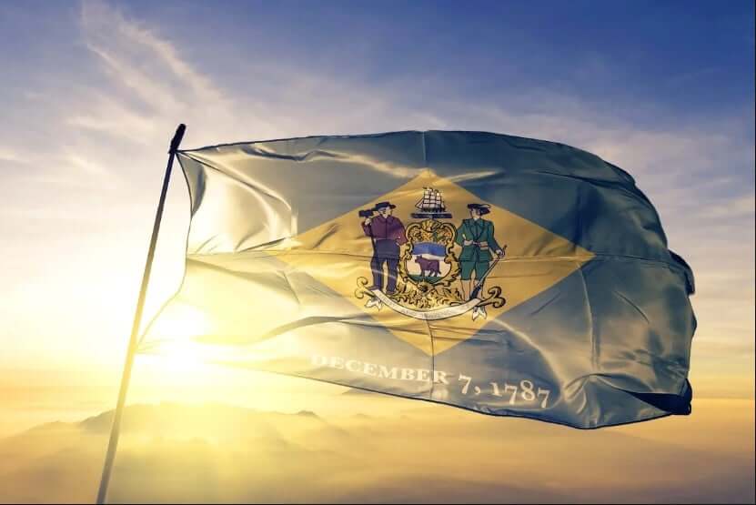 The Delaware state flag waves against a scenic sunrise backdrop, featuring the state coat of arms on a buff diamond background and the date "December 7, 1787" below.