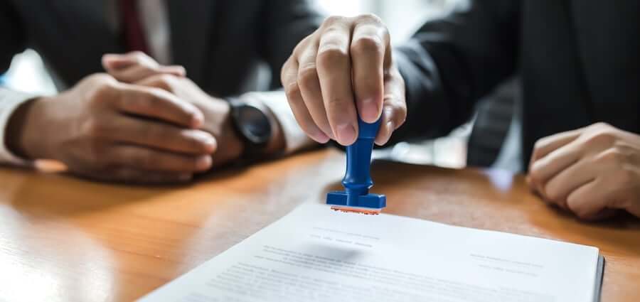 Two people in business attire are at a desk, with one person's hand pressing a blue stamp onto a document. The focus is on the stamping action, while the other person's hands are folded on the table.
