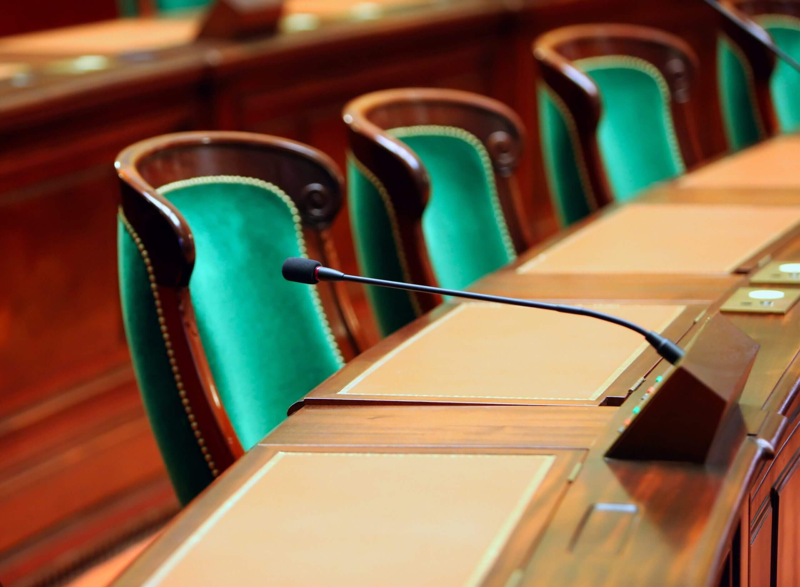 A row of empty wooden desks with green upholstered chairs in a formal meeting room. Each desk has a microphone and light brown leather writing pads. The room has an elegant and professional atmosphere.