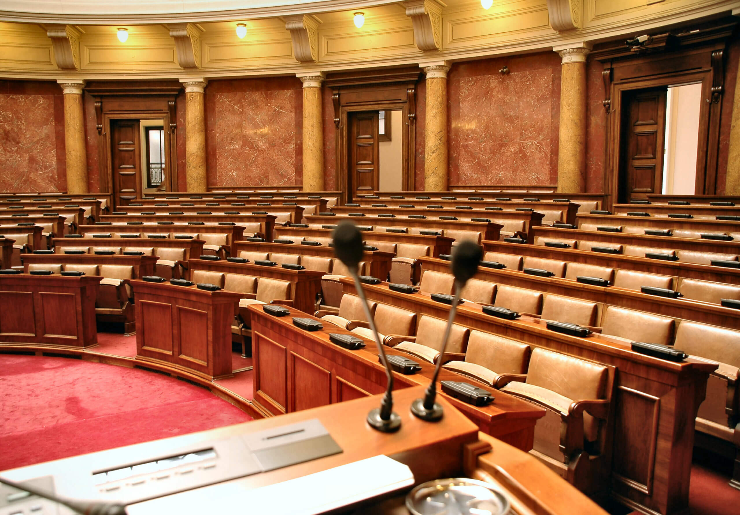 An empty parliamentary chamber with rows of wooden desks and beige leather chairs. Two microphones are visible on a podium in the foreground. The walls are adorned with marble and columns, and a curved ceiling holds several light fixtures.