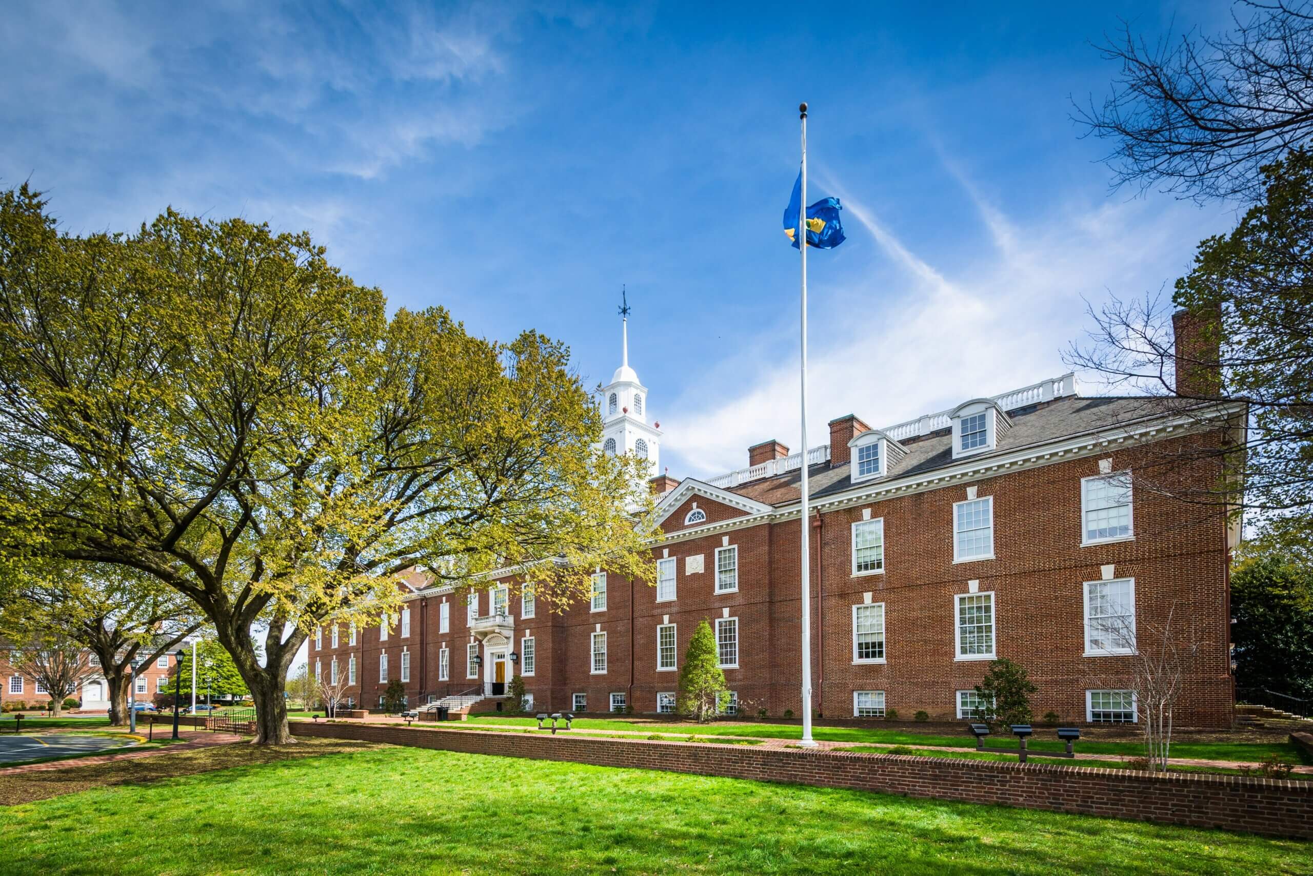 A brick university building stands under a bright blue sky, adorned with a central cupola. A flag flies on a tall pole in front of the building. Leafy trees frame the scene, casting gentle shadows on the green lawn.