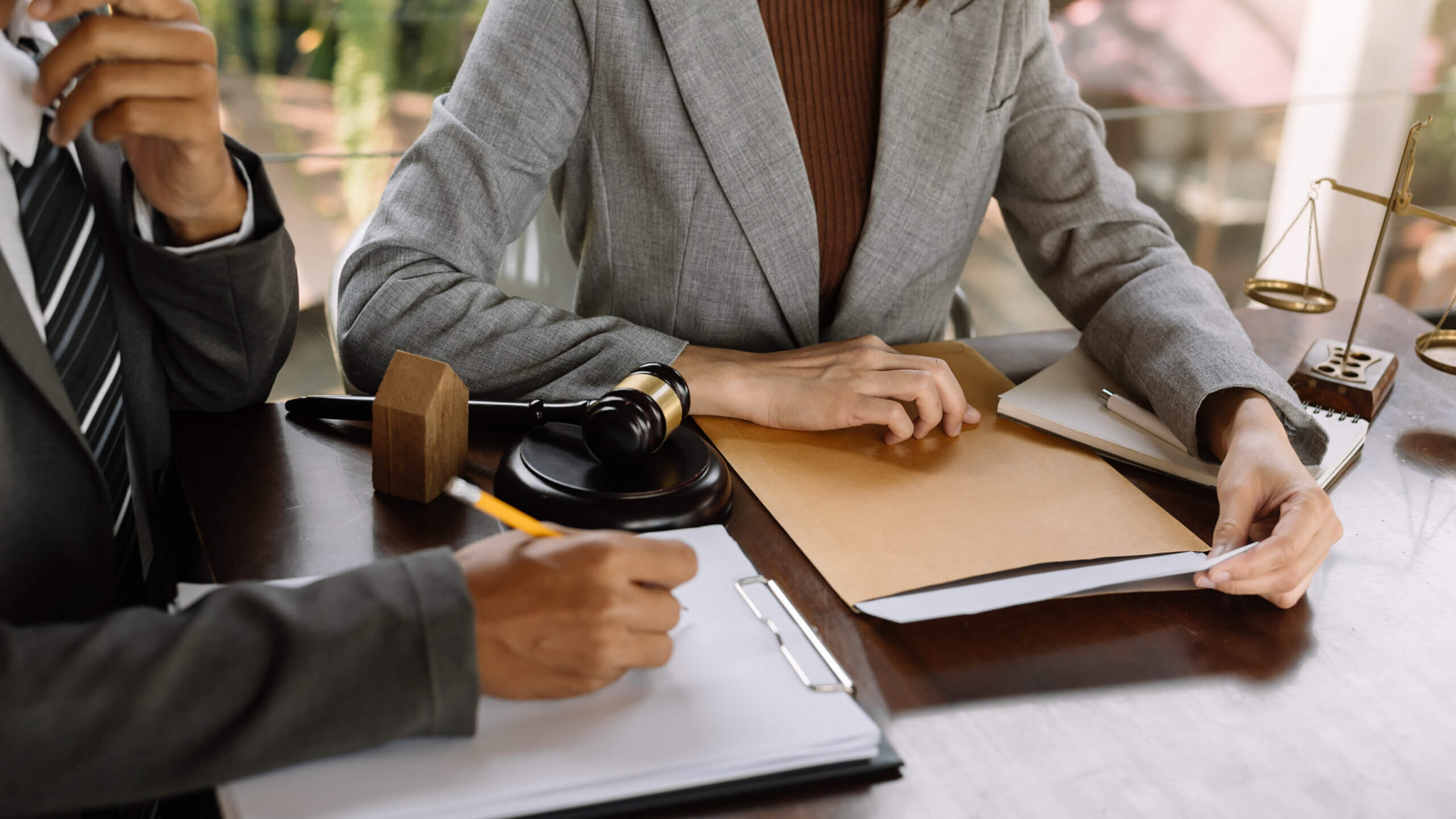 Two people in professional attire sit at a wooden table. One holds a pen over a clipboard, while the other sits with a folder. A gavel and scales of justice are on the table, suggesting a legal setting.