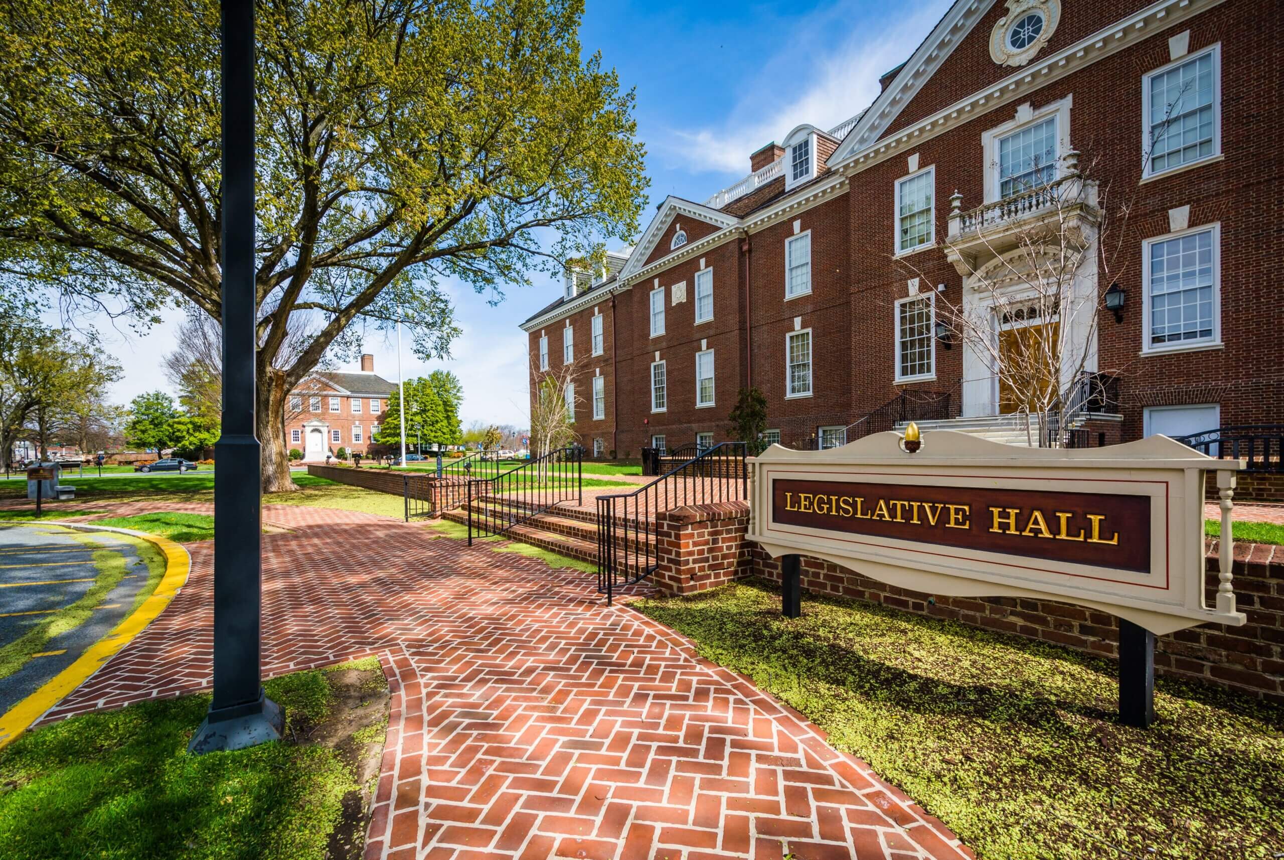 A red-brick path leads to a large red-brick building named Legislative Hall. The scene features green lawns, a tree to the left, and a clear blue sky. Another similar building is visible in the background.