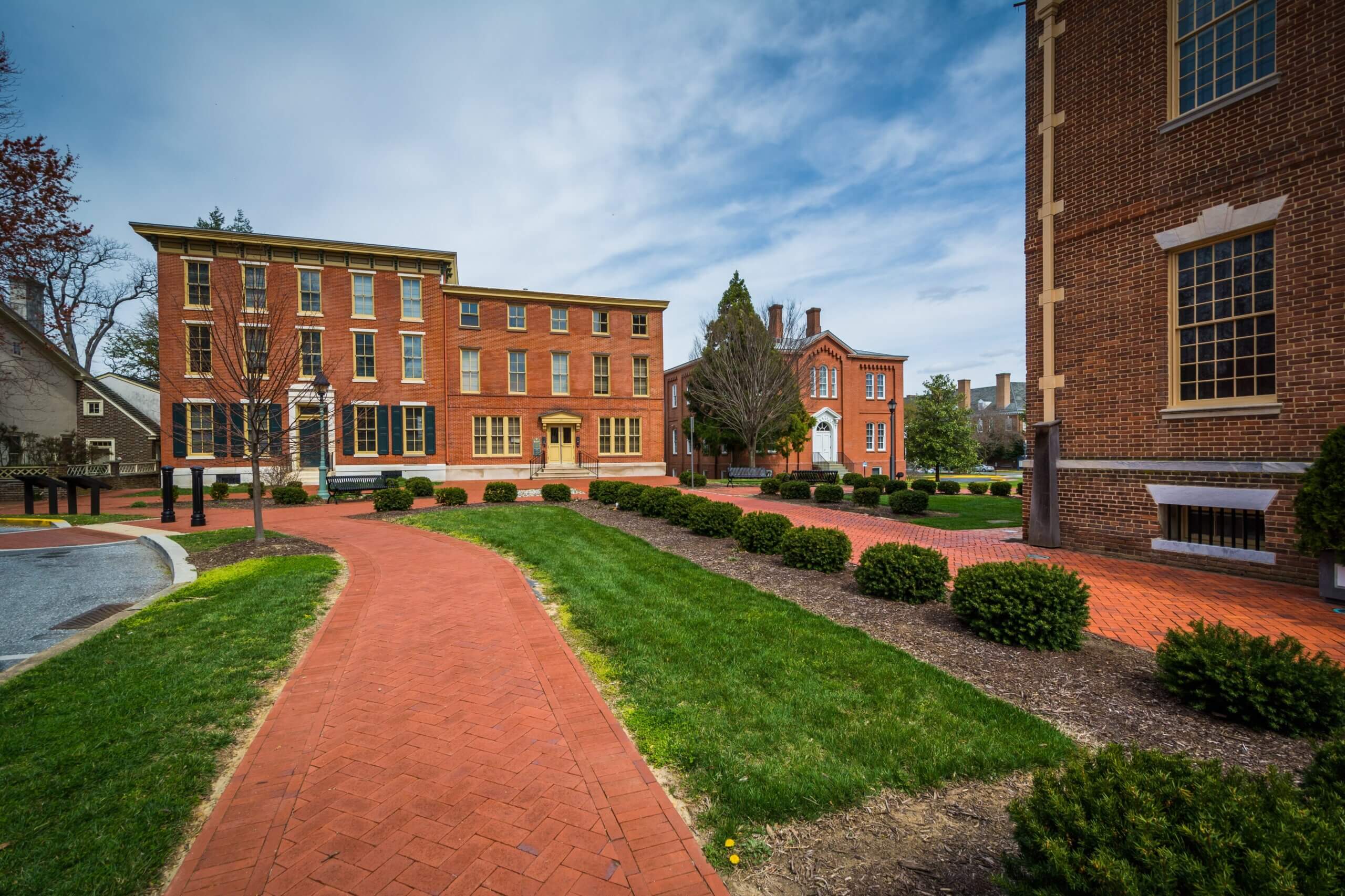 A historic site featuring brick buildings, a red brick walkway, and manicured lawns with green grass and shrubs. The scene is bright and clear under a partly cloudy sky.
