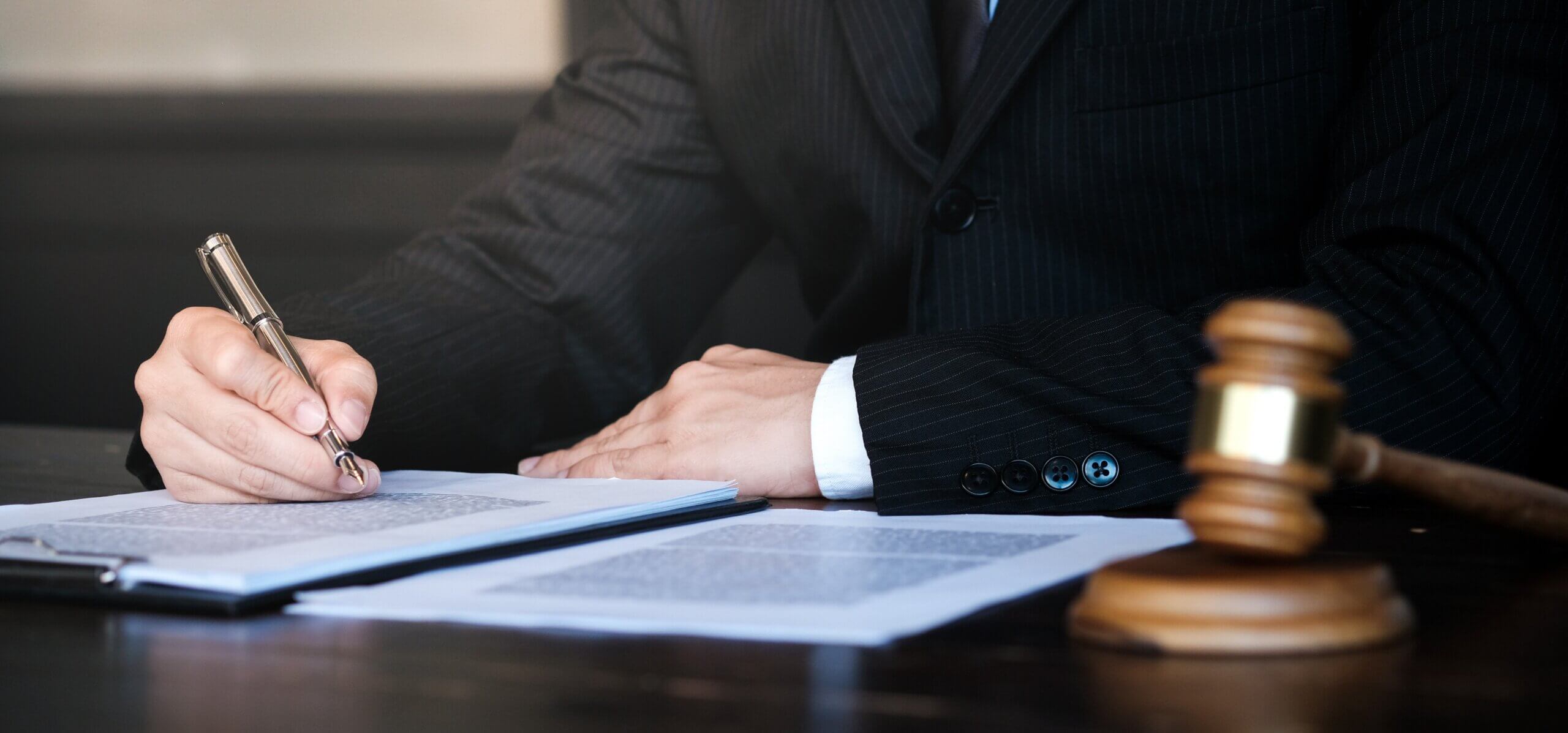 A person in a suit sits at a desk, holding a pen and writing on documents. A gavel is placed nearby on the desk, suggesting a legal or judicial setting. The background is blurred.