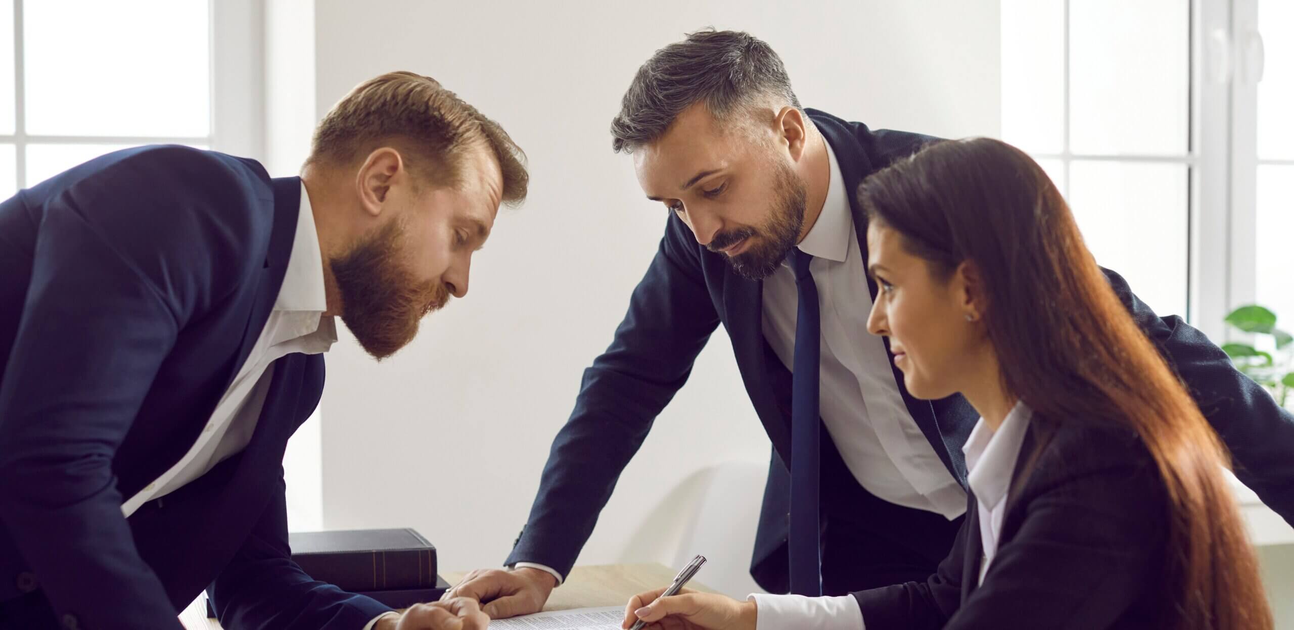 Three people in business attire discussing documents related to special agency representation at a table in an office. One woman is writing on a paper while two men stand and converse. A gavel is visible on the table.