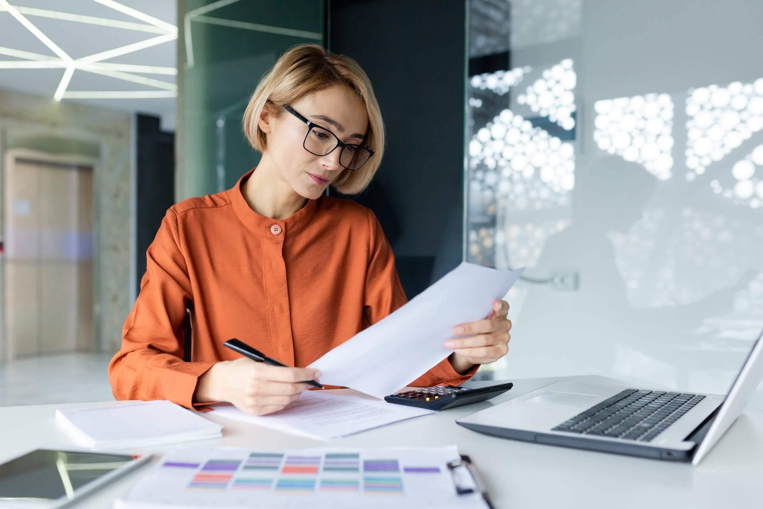 A person with short hair, wearing a rust-colored shirt and glasses, sits at a desk reviewing documents. A laptop, calculator, and colorful charts are on the table in a modern office space.