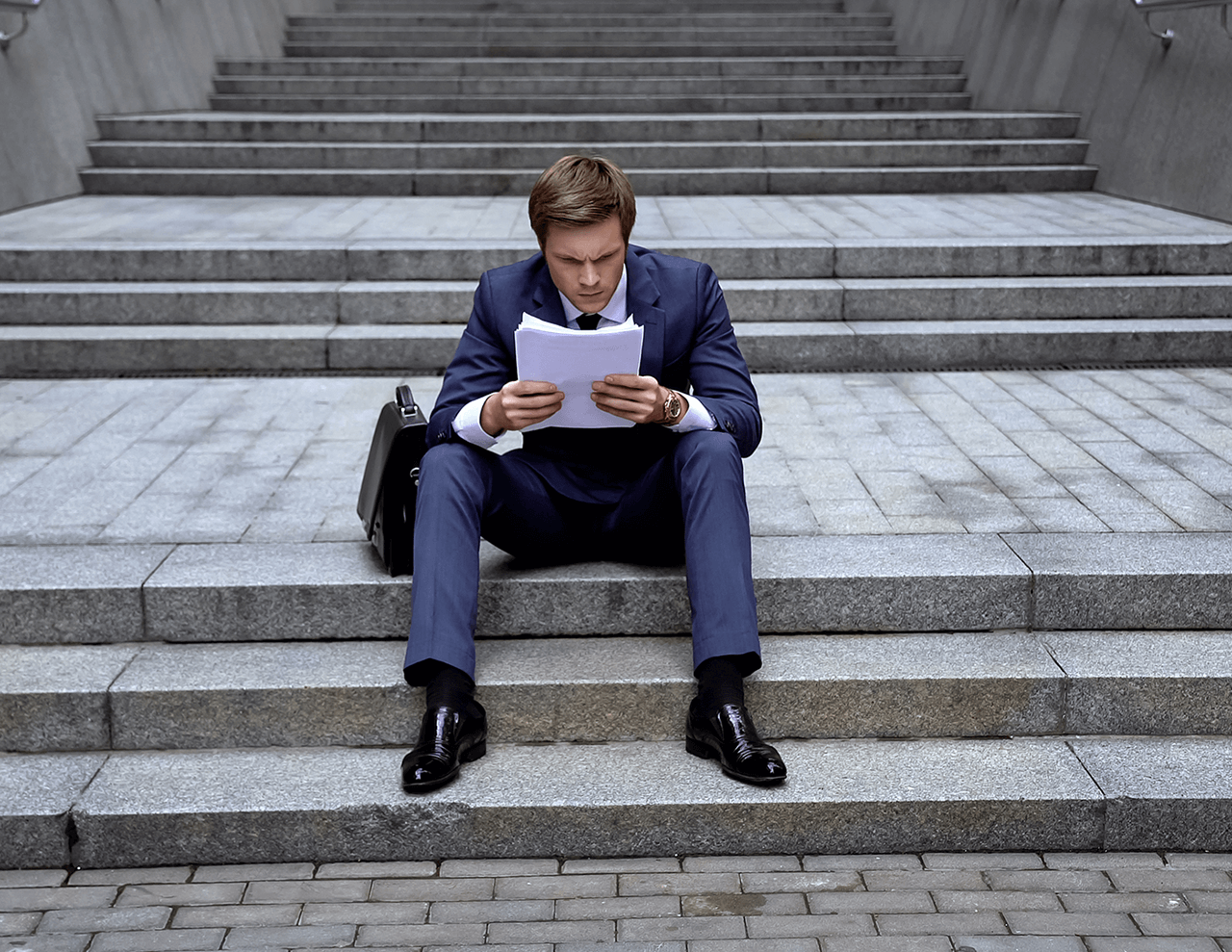 A man in a suit sits on outdoor steps, intently reading a stack of papers. A briefcase is beside him. The background shows multiple stone steps leading upwards.