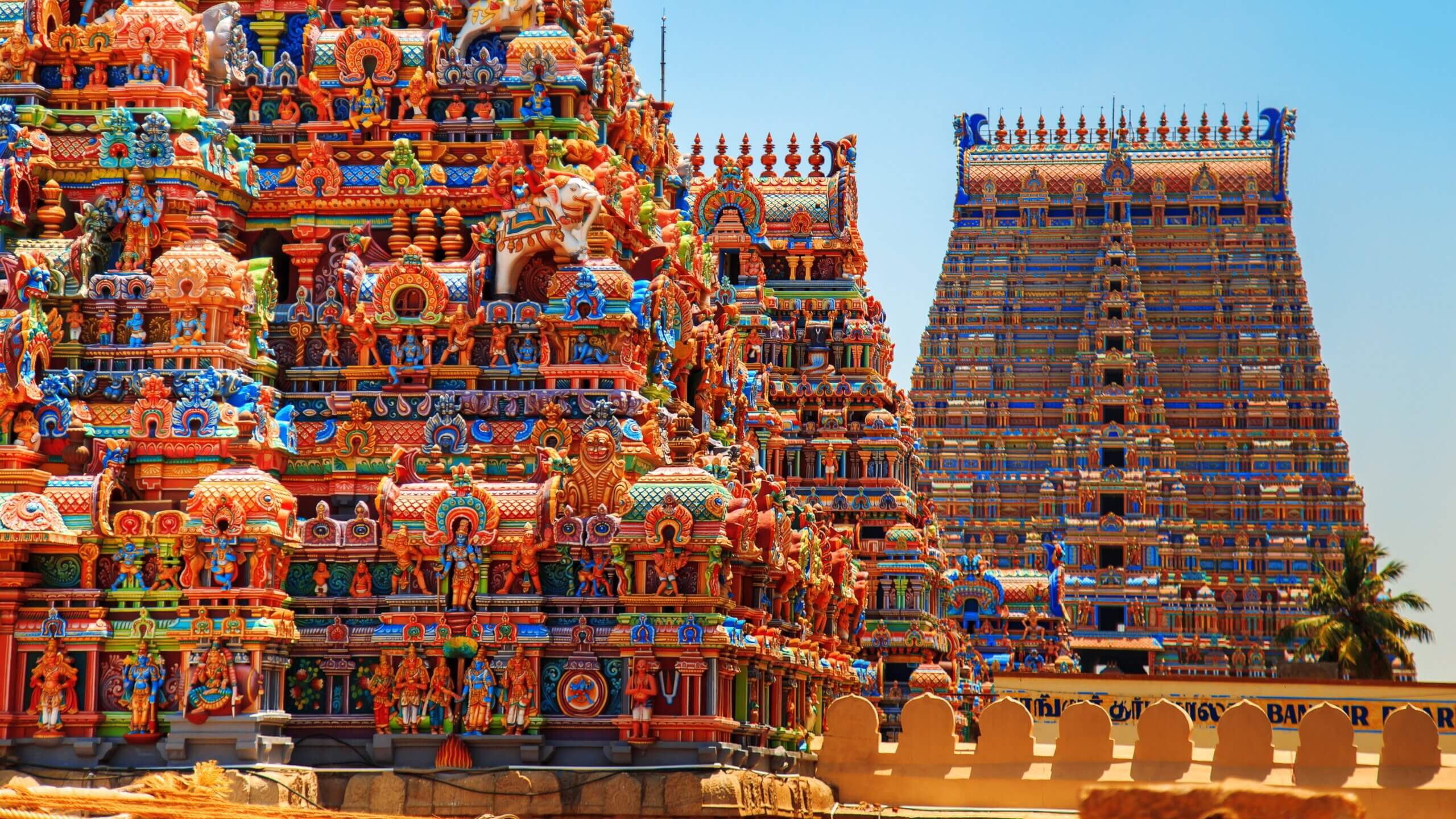 Vibrant, intricately decorated towers of a Hindu temple, featuring detailed sculptures and colorful carvings against a clear blue sky. A larger tower is visible in the background, partially obscured by the foreground structures.
