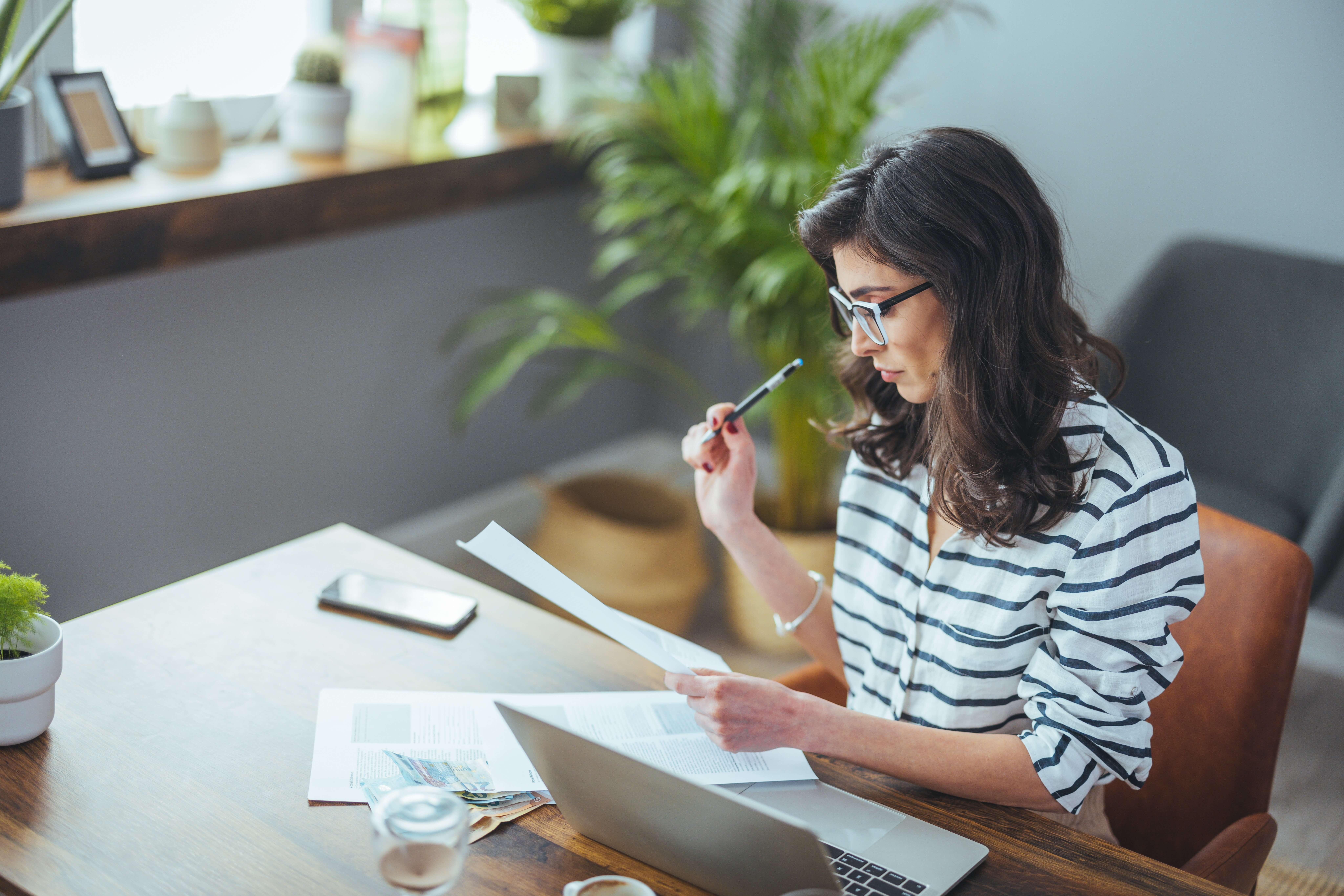 A woman with glasses sits at a wooden desk in a home office, holding a pen and reading documents. A laptop, phone, and cup are on the table. The room is decorated with plants and has a soft lighting ambiance.