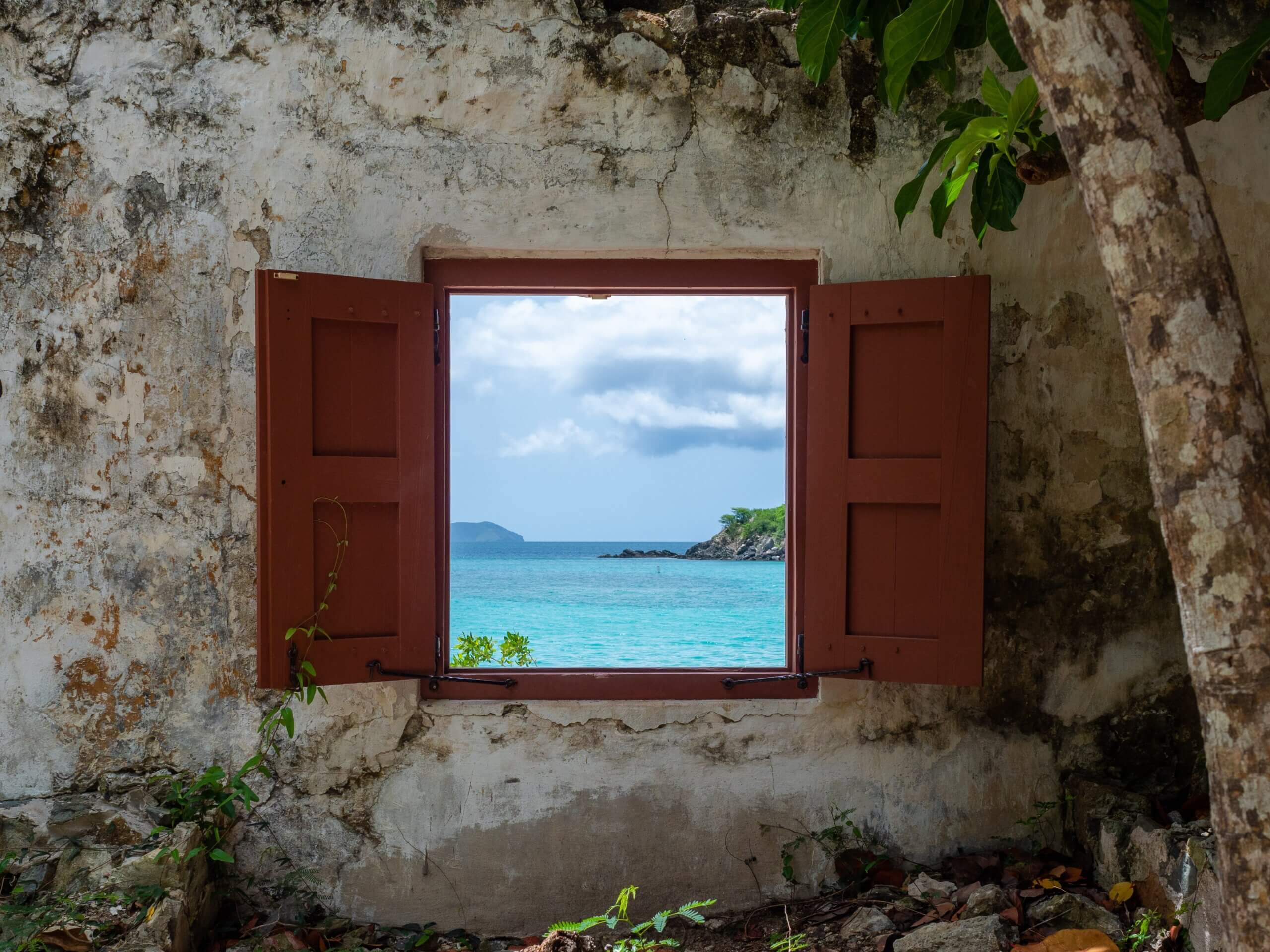 A view of the ocean through an open window with brown wooden shutters framed in an old, weathered stone wall. Lush greenery partially surrounds the scene, and a tree trunk is visible to the right. The sky is cloudy above the turquoise sea.