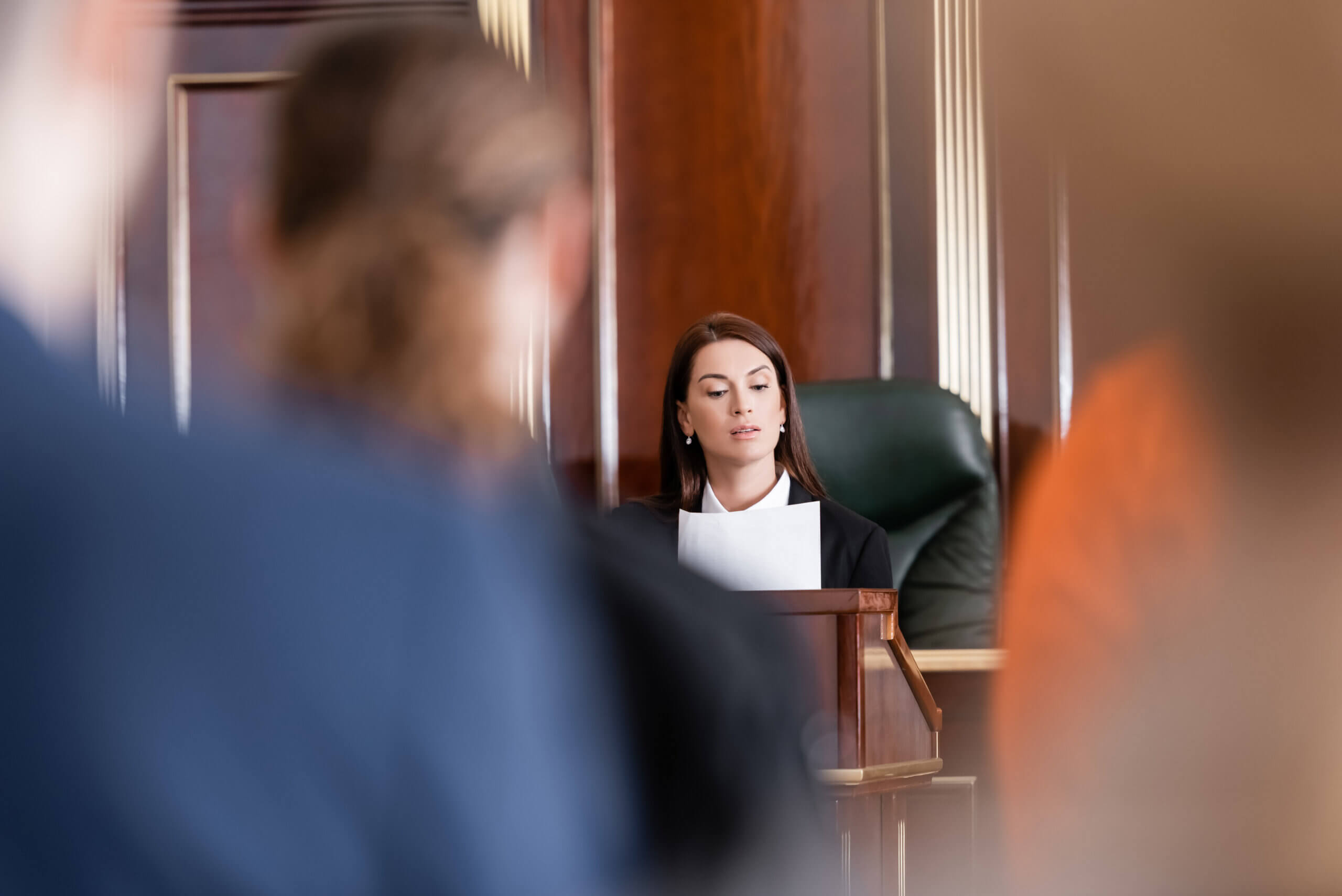 A judge in a courtroom is reading from a paper, seated in a high-backed chair. The room has wood paneling and people are blurred in the foreground. The scene conveys a formal legal setting.