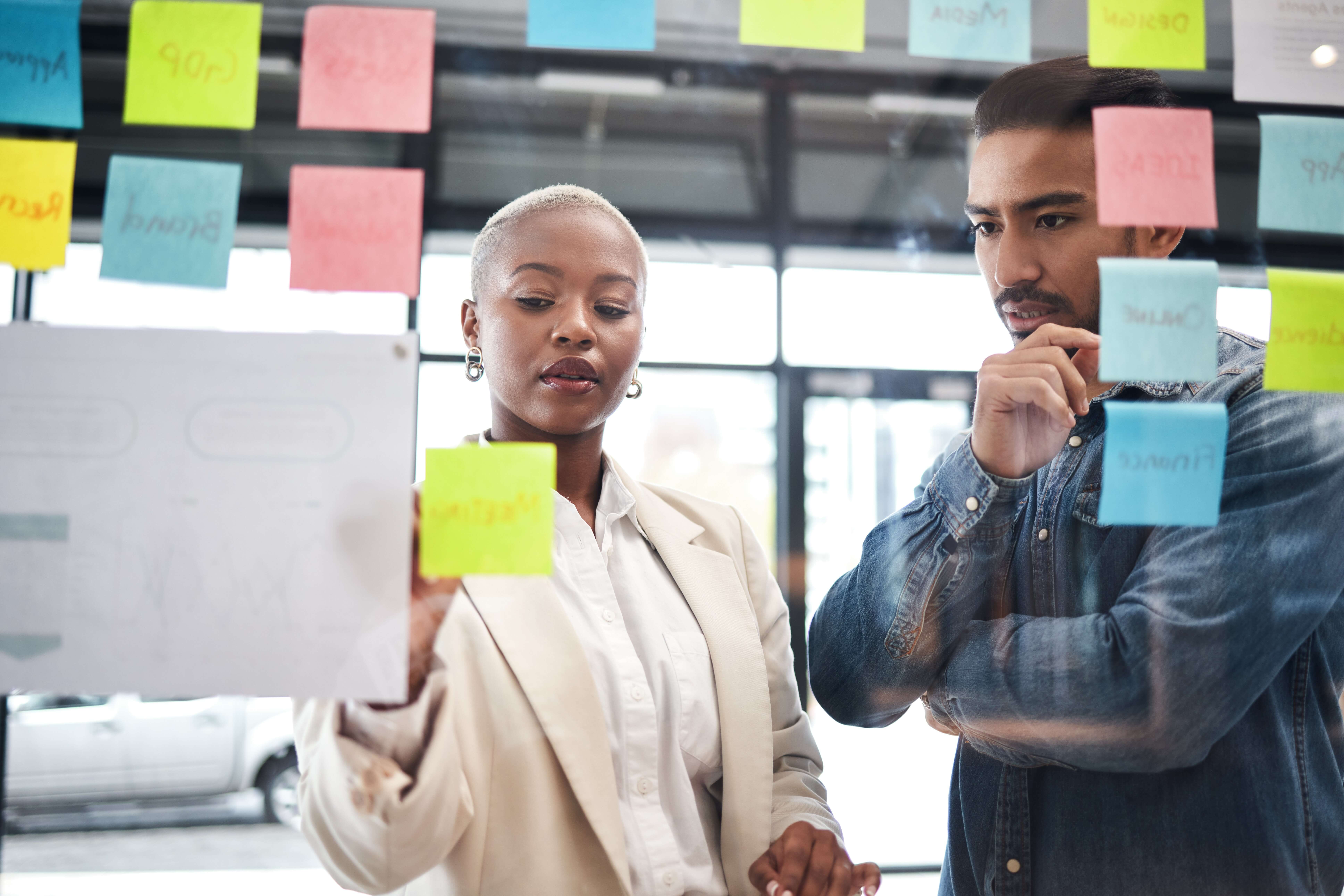 A woman and a man stand in front of a glass wall covered with colorful sticky notes. The woman is pointing at a note, and the man is observing thoughtfully. They appear to be in a modern office setting, focused and engaged.