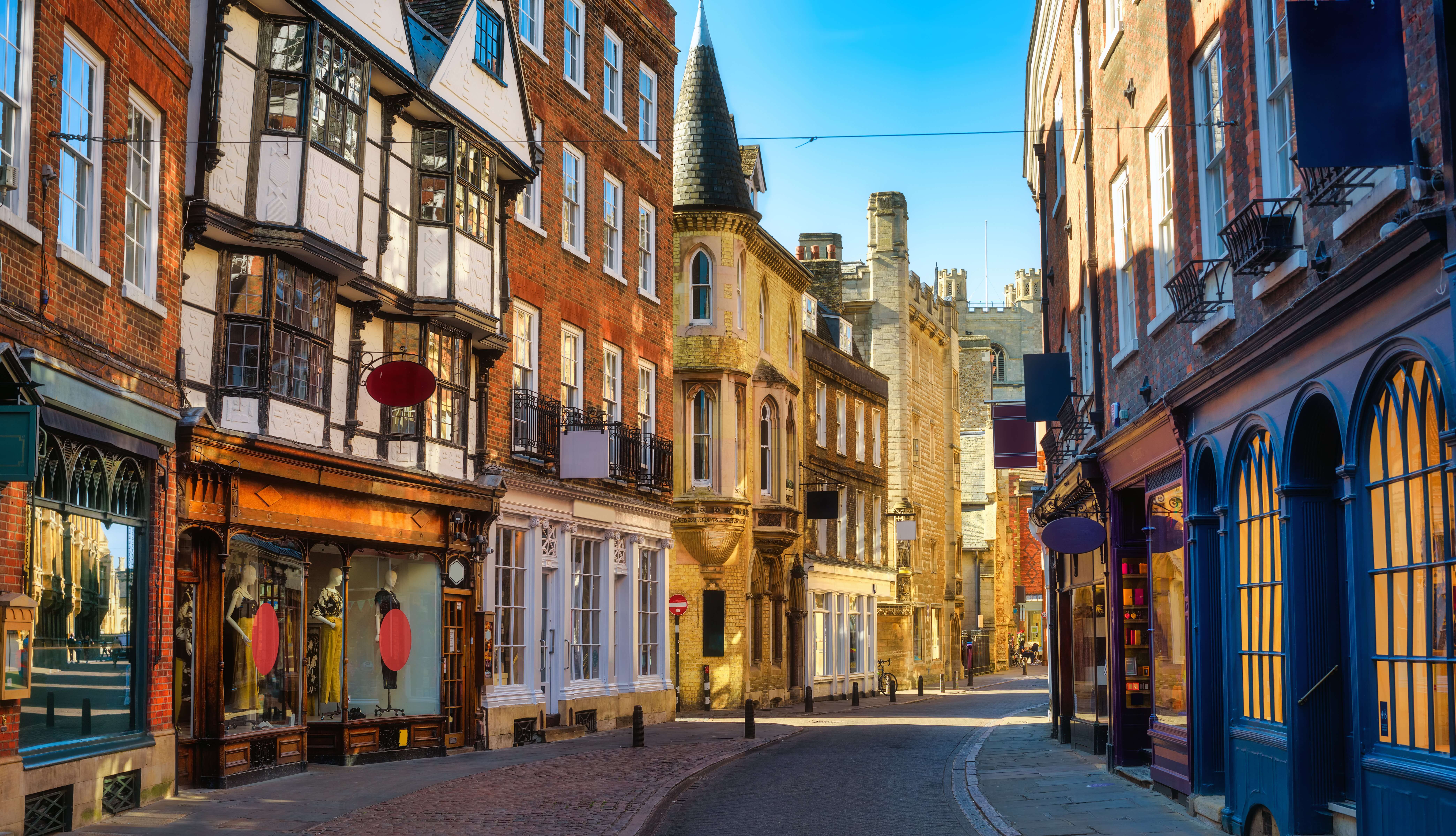 A picturesque narrow street lined with historic brick and timber-framed buildings under a clear blue sky. The street is paved with cobblestones and reflects warm sunlight, creating an inviting and charming atmosphere.