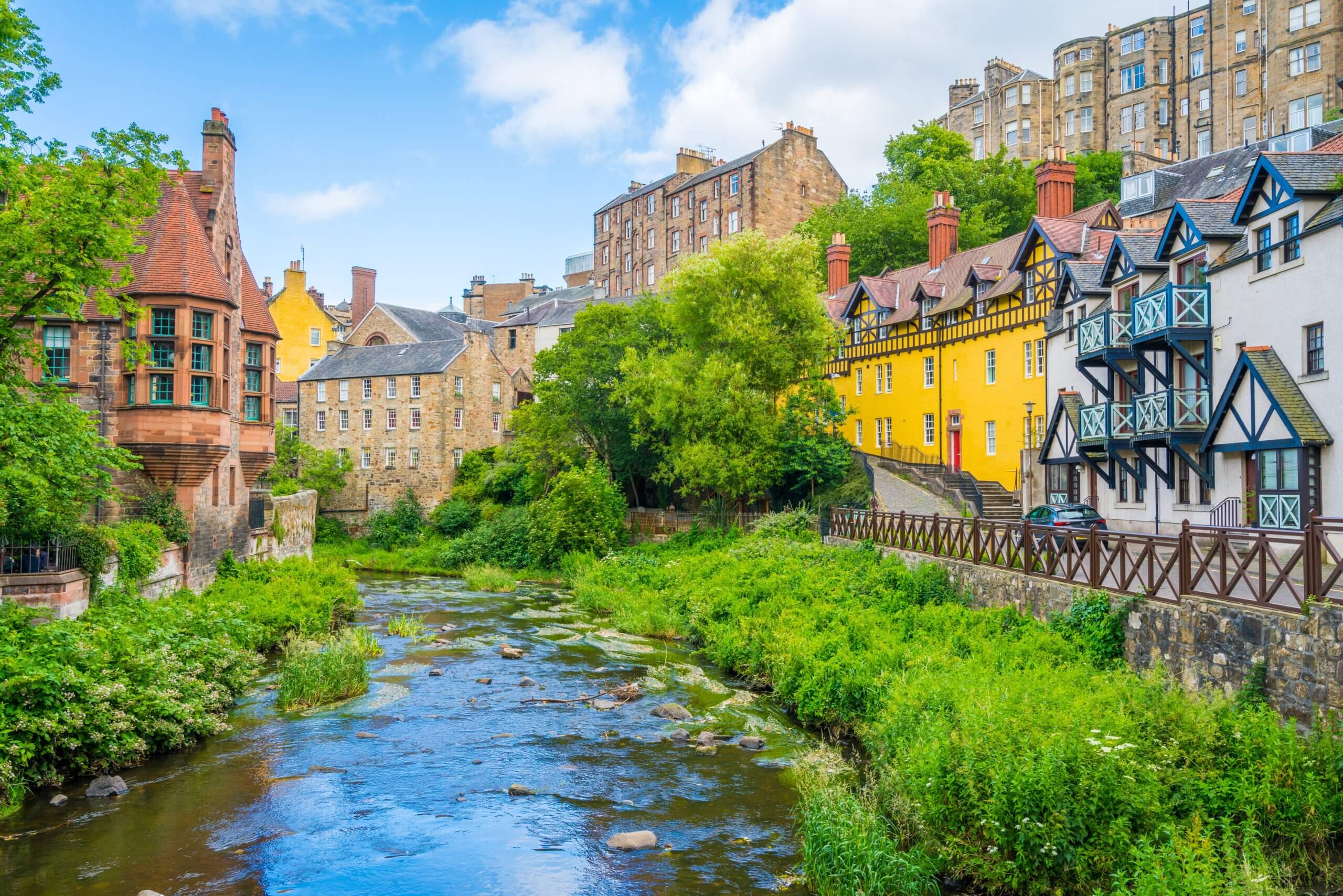 Colorful buildings line a lush riverbank in a quaint village. A small stream flows through, surrounded by greenery. The sky is blue with few clouds, and the scene has a charming, tranquil atmosphere.