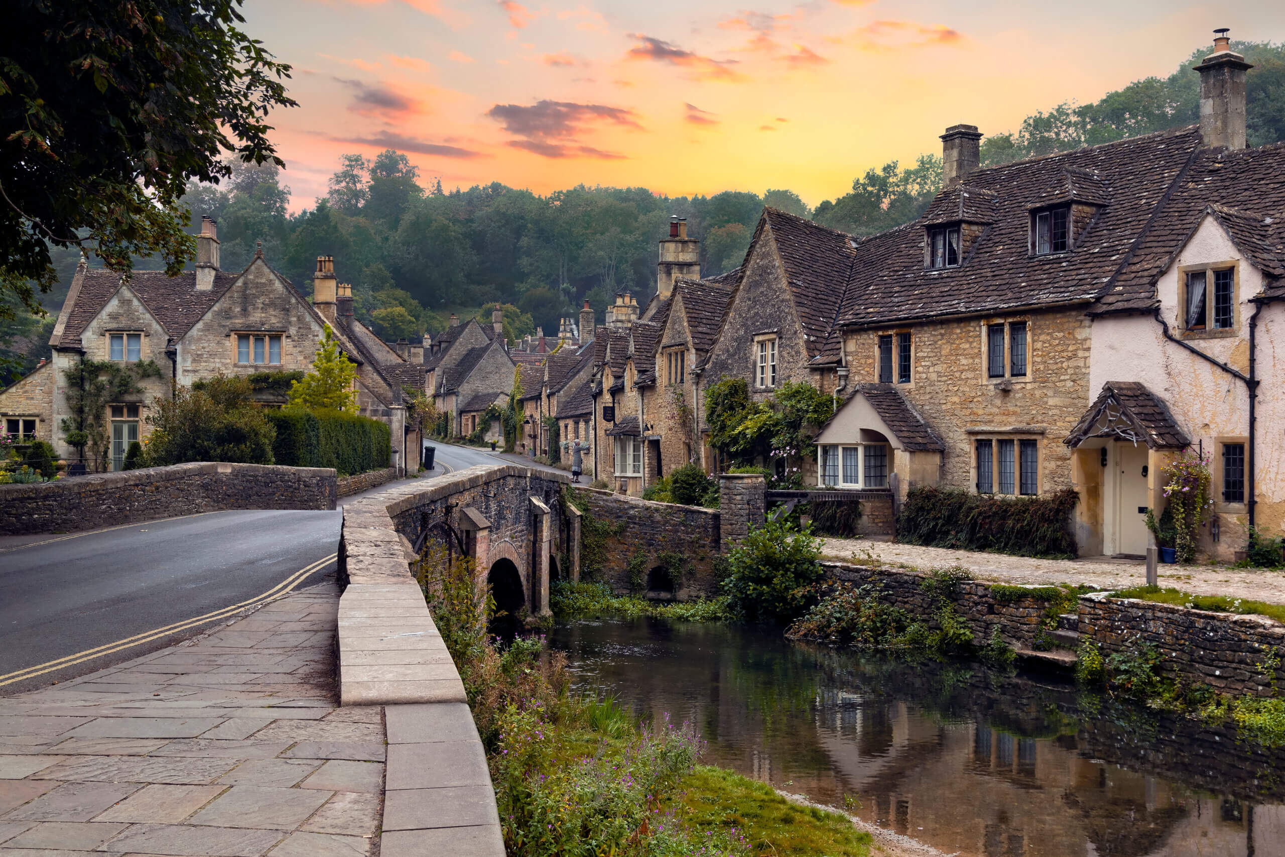 Quaint village scene at sunset with stone cottages lining a curved road and a narrow stone bridge over a calm river. Lush greenery surrounds the area, as warm light from the setting sun casts a gentle glow over the picturesque landscape.