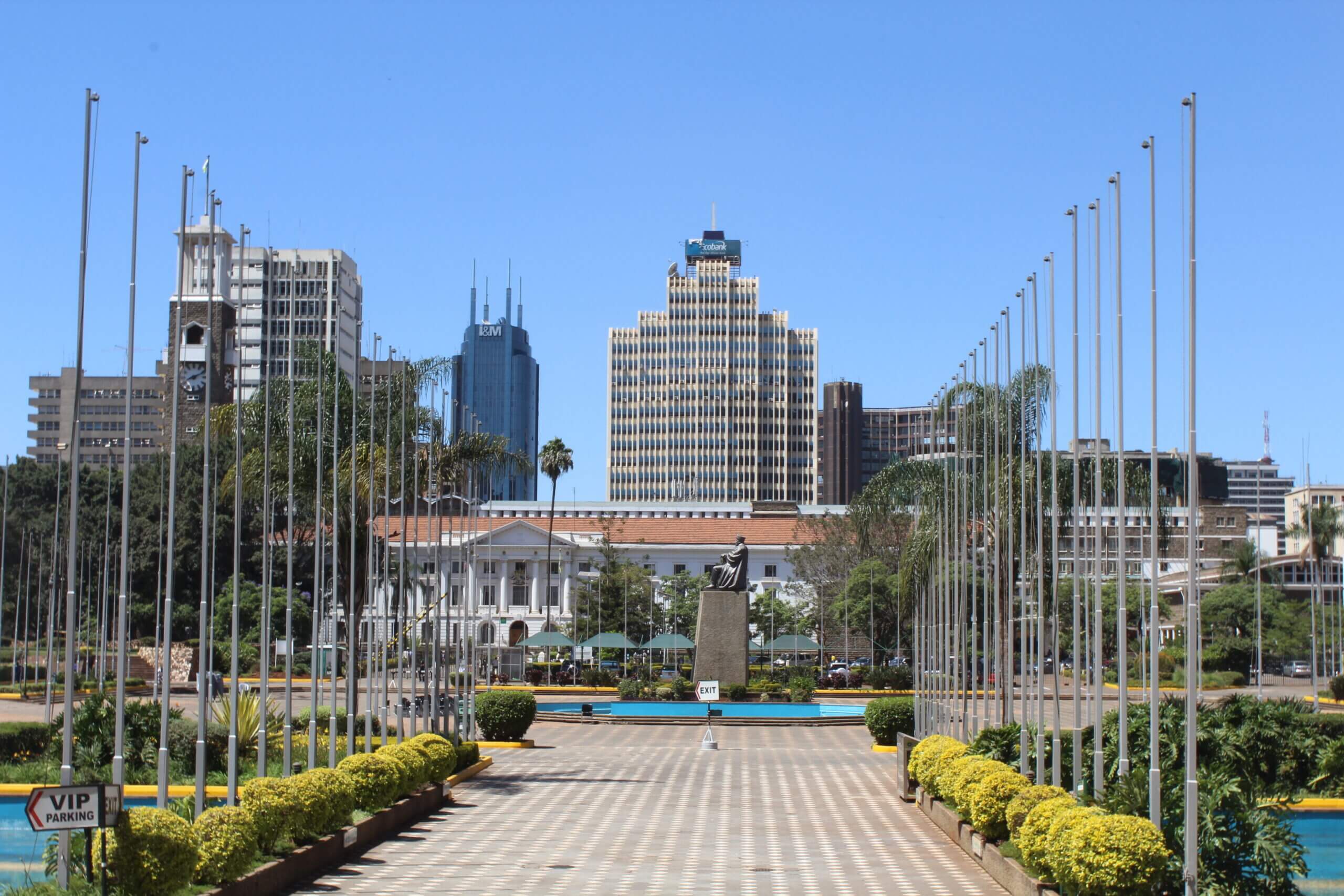 A view down a wide path lined with flagpoles leads to a statue and a large building with a clock tower, surrounded by modern skyscrapers. The sky is clear and blue. Green hedges and neatly trimmed bushes border the path.